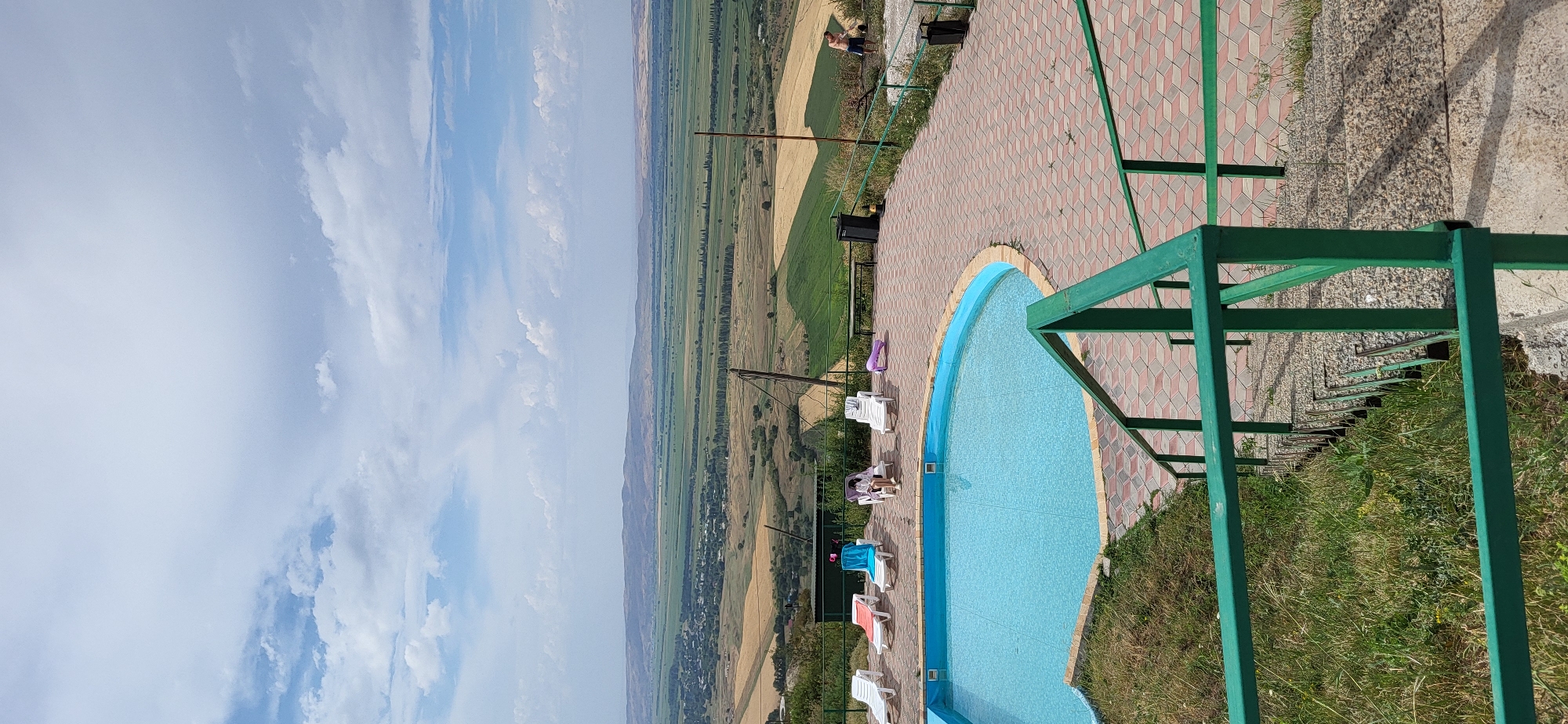 The image depicts an outdoor swimming pool area with a panoramic view of a valley. The pool is kidney-shaped and filled with bright blue water, surrounded by a paved patio made of reddish-brown and grey interlocking bricks. Several white lounge chairs are arranged around the pool, with towels draped on some of them. One person is seated on a lounge chair, wearing a white robe. Further to the right, another person is visible standing near a dark green trash bin. In the distance, a vast valley unfolds, characterized by agricultural fields of varying colors, including green and golden hues, and scattered patches of trees. Rolling hills form the horizon under a sky with scattered white clouds. To the left and in the foreground, a set of outdoor stairs with a green metal railing descends from the pool area. Green grass and vegetation grow on the embankment next to the stairs. A person in blue shorts and a bare chest is seen in the far right background of the image, appearing to be outdoors. The location is identified as Syrymbet, Kazakhstan.