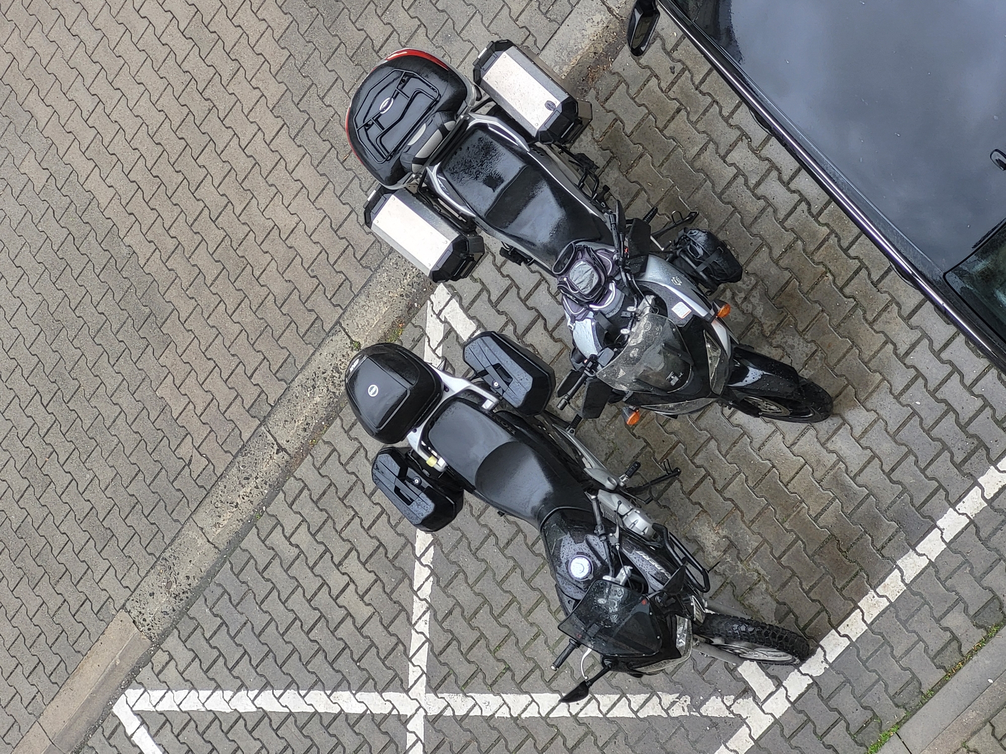The image depicts two motorcycles parked in a herringbone brick parking space, with the corner of a black car visible in the upper right. The scene suggests an urban environment, though the specific location within Unknown, Unknown cannot be determined. Both motorcycles appear to be equipped for touring, featuring luggage boxes or panniers. One motorcycle has silver panniers and a red-accented top box, while the other has smaller, black luggage boxes. The parking space is marked by white lines. The motorcycles are wet, indicating recent rain. There is no visible human activity.