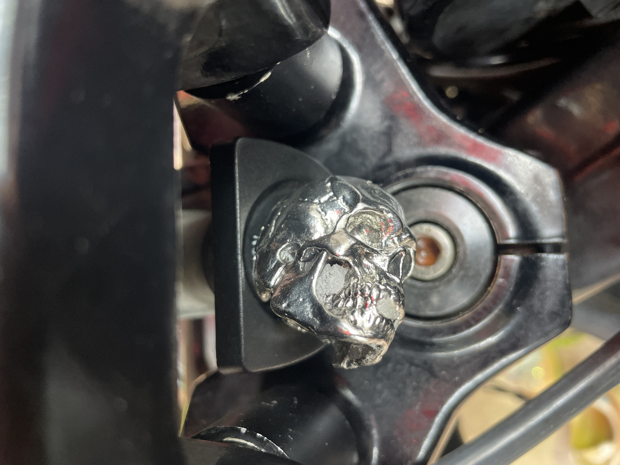 The image depicts a close-up of a motorcycle's front end. A prominent feature is a chrome-colored skull decoration attached to the motorcycle's fork clamp. The motorcycle is black, with some red reflections visible. The environment is out of focus, but there is a hint of yellow and another vehicle in the background. Based on the prompt, this location is in Thep Krasatti, Thailand.