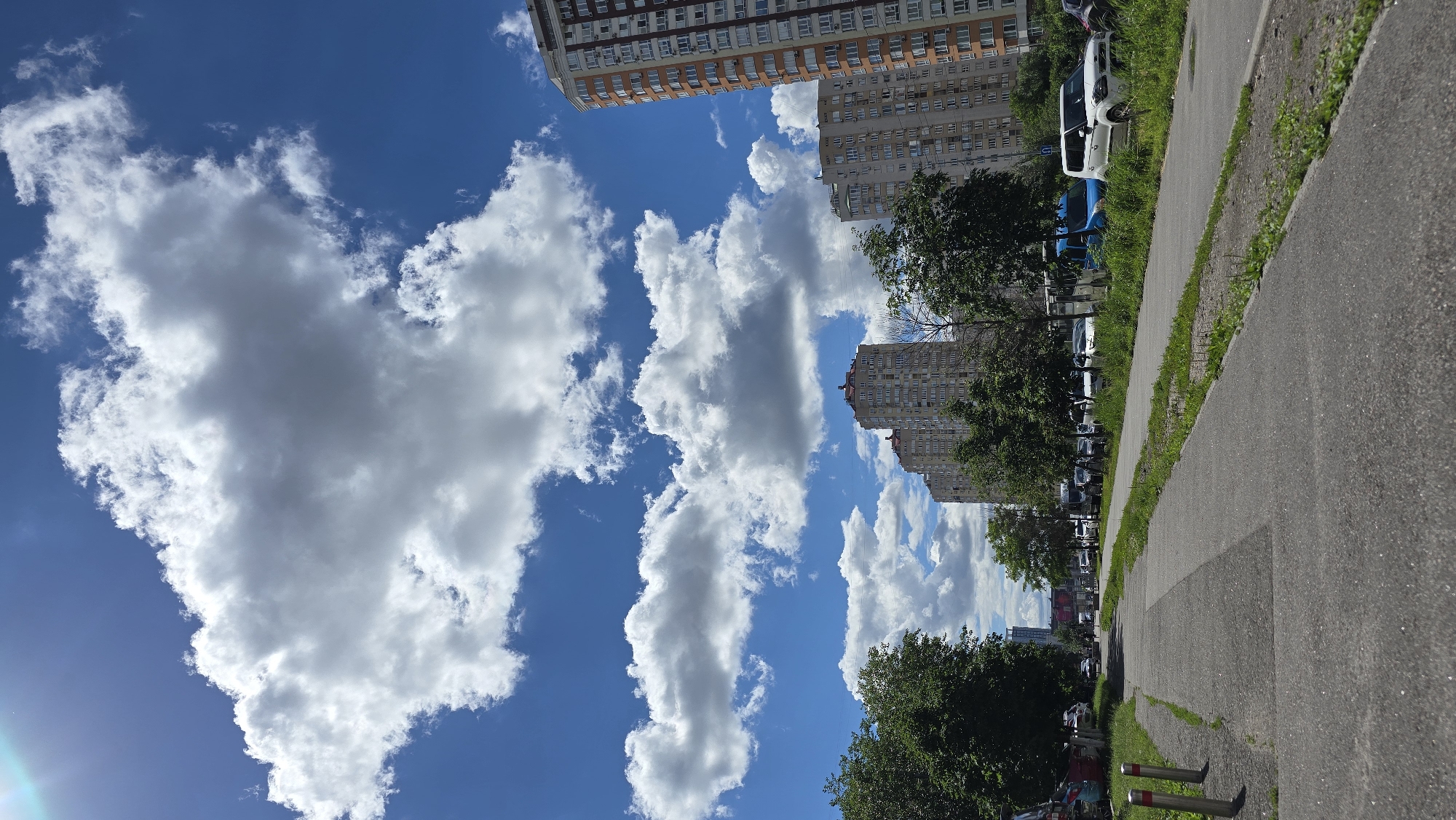 This is a vertical, eye-level, full outdoor shot of a residential street in Kyiv, Ukraine. The scene is captured on a bright, sunny day with a clear blue sky dotted with large, fluffy white clouds. A paved walkway or bike path runs diagonally from the lower left foreground towards the center right. On the left side of the walkway, there is a strip of green grass and a few cylindrical metal bollards. The right side of the walkway is bordered by more grass and a road lined with parked cars.
In the background, several multi-story residential buildings are visible. The building on the far right is taller and has an orange-colored facade with white windows and balconies. To its left, a series of slightly shorter, lighter-colored buildings with numerous windows stretch across the frame. Trees with dense green foliage are scattered in front of and between the buildings, some tall and slender, others shorter and bushier. The road appears to be a street with several cars parked along its side, including a white SUV, a blue van, and various other vehicles. The environment appears to be a typical urban residential area. There are no people visible in the frame.