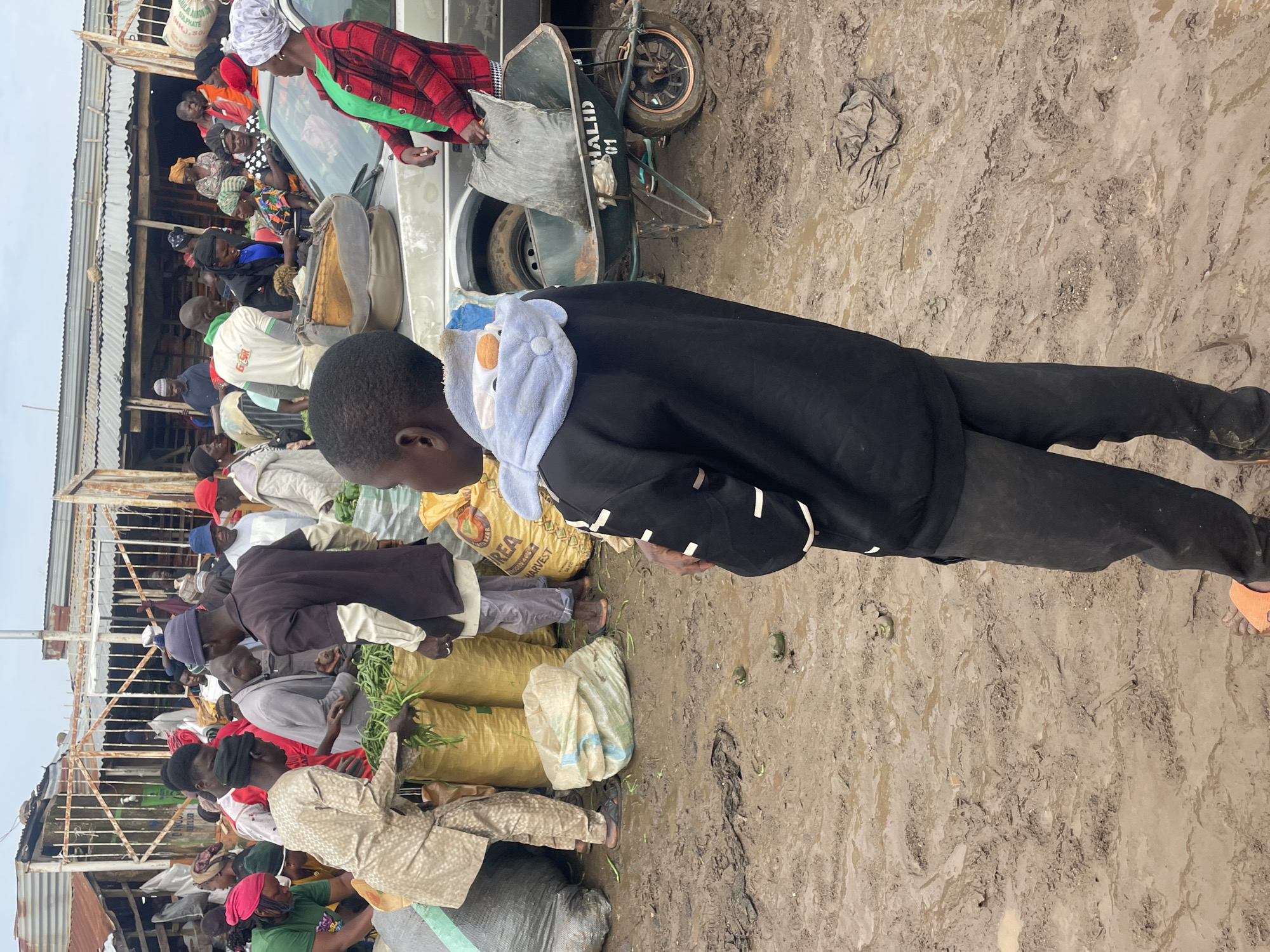 The image depicts a bustling outdoor market scene in Gura Top, Nigeria. The ground is muddy and wet, indicating recent rain. Numerous people are gathered, engaged in various activities related to commerce. In the foreground, a young boy stands with his back to the camera, wearing a black hoodie with a light blue hood and dark pants.
Various vendors are seen interacting with customers and handling goods. Large sacks, likely containing produce or grains, are prominent throughout the scene. One man in a dark brown shirt and a blue cap is handing green vegetables to another person. A woman in a red and black checkered jacket is near a silver car. A wheelbarrow is visible, holding a black bag. The market appears to be set up with makeshift stalls and structures, some with corrugated metal roofing. The overall atmosphere suggests a busy marketplace in full swing.