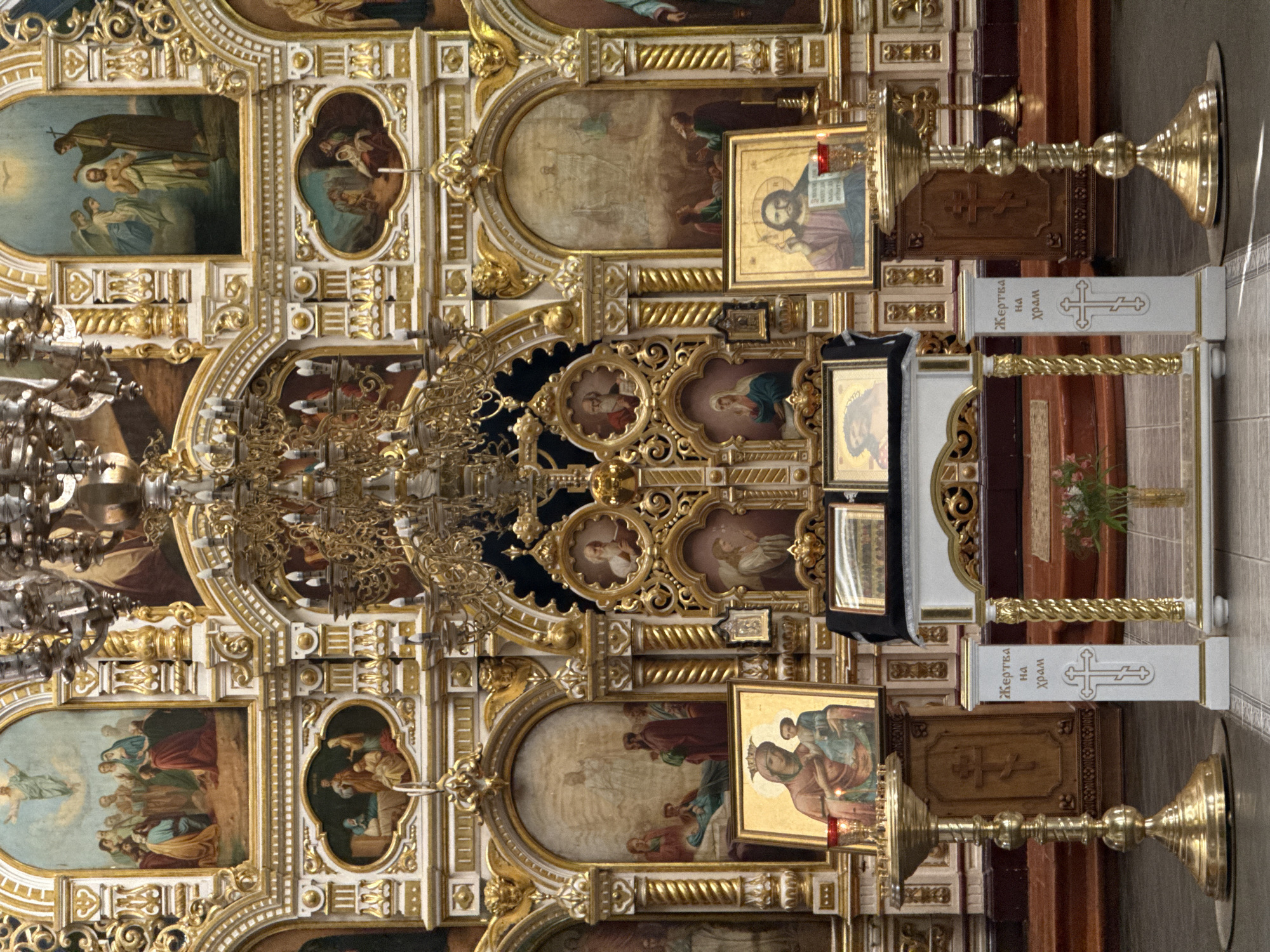 The image depicts a richly decorated interior, likely within a religious building in Votkinsk, Russia. Specifically, it appears to be an Orthodox church, judging by the iconography and architectural details. The scene showcases an ornate altar or iconostasis, featuring multiple layers of gilded frames, paintings, and religious icons. A large chandelier hangs from the ceiling, adding to the grandeur of the setting. In the foreground, a candle stand is visible. The overall impression is one of opulence and religious reverence.