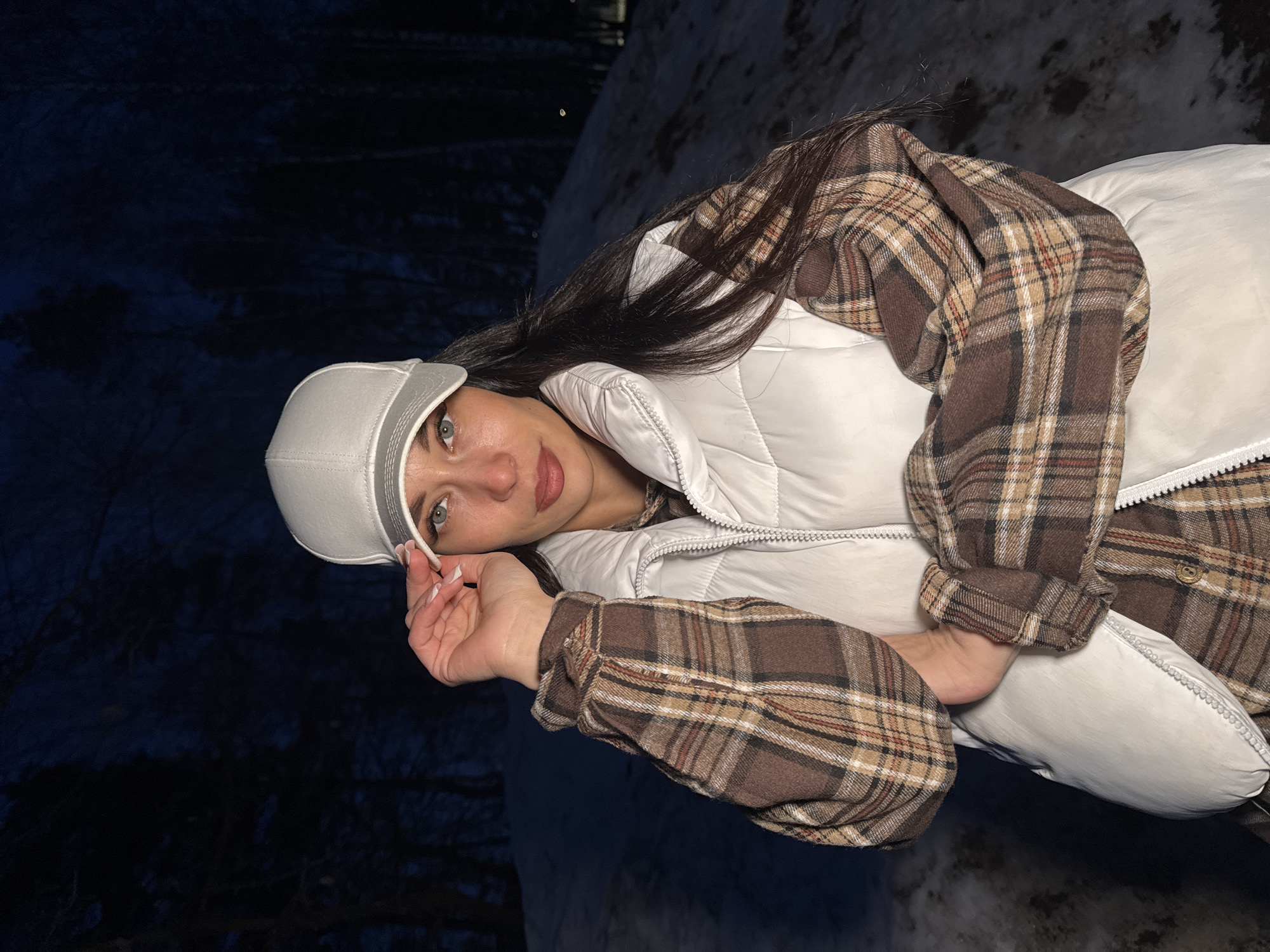 A young woman with long dark hair and light eyes is posing for a photo outdoors. She is wearing a white baseball cap, a white puffy vest, and a brown and tan plaid shirt. Her left hand is raised near her face.
The background is dark, suggesting it is night or a dimly lit area. Patches of snow are visible on the ground. Based on the visible details and the location provided (Chaikovsky, Russia), the environment seems to be an urban park or green space during the winter.