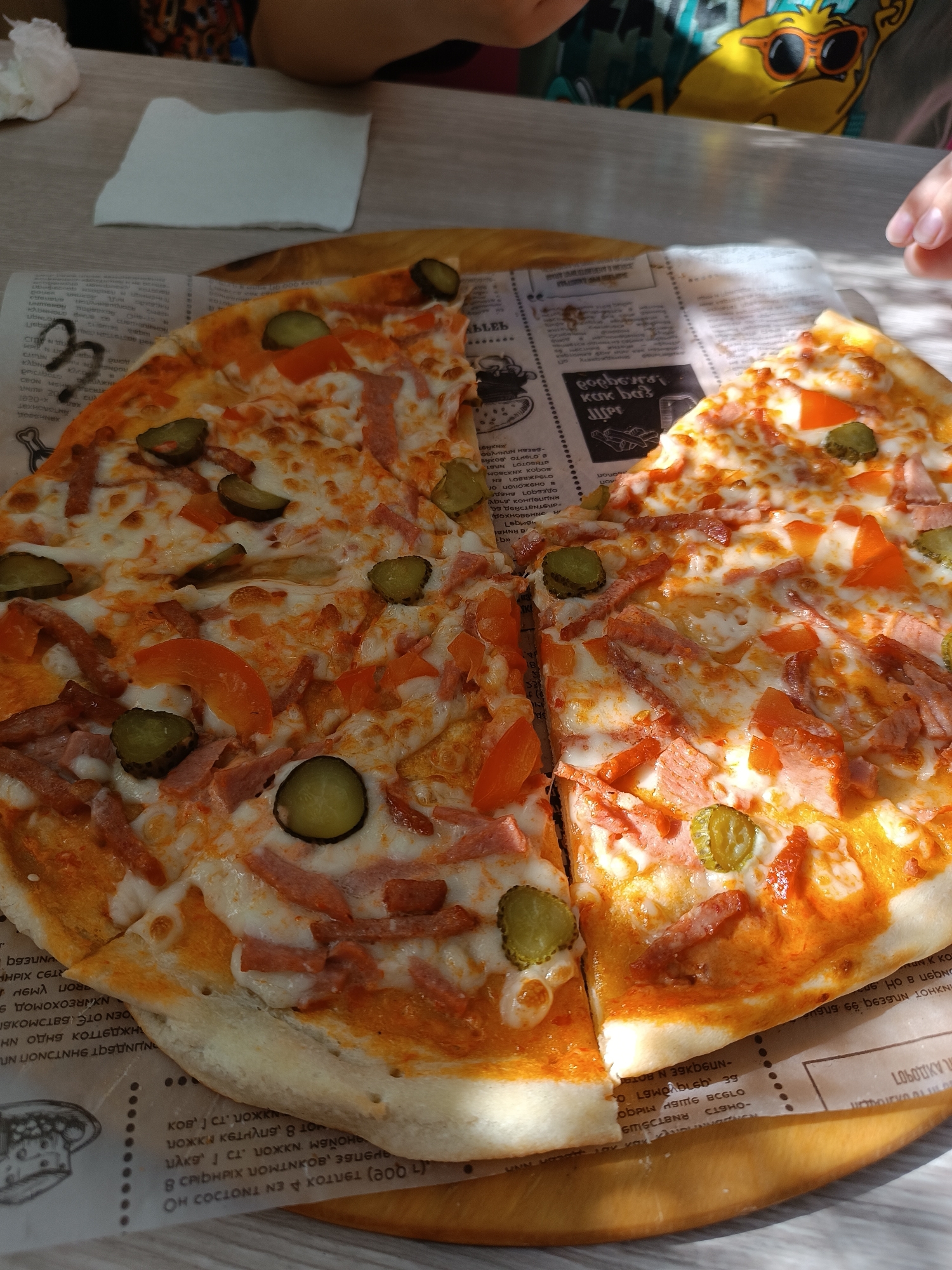 The image depicts a partially eaten pizza on a wooden serving board covered with a newspaper-style printed paper. The pizza toppings include cheese, a tomato-based sauce, sliced pickles, diced tomatoes, and processed meat strips. Two people are partially visible at the top of the frame, one wearing a colorful patterned shirt. The pizza is on a table indoors, with a white napkin nearby. The newspaper-style paper has text in Russian, indicating the location may be in Chaikovsky, Russia.