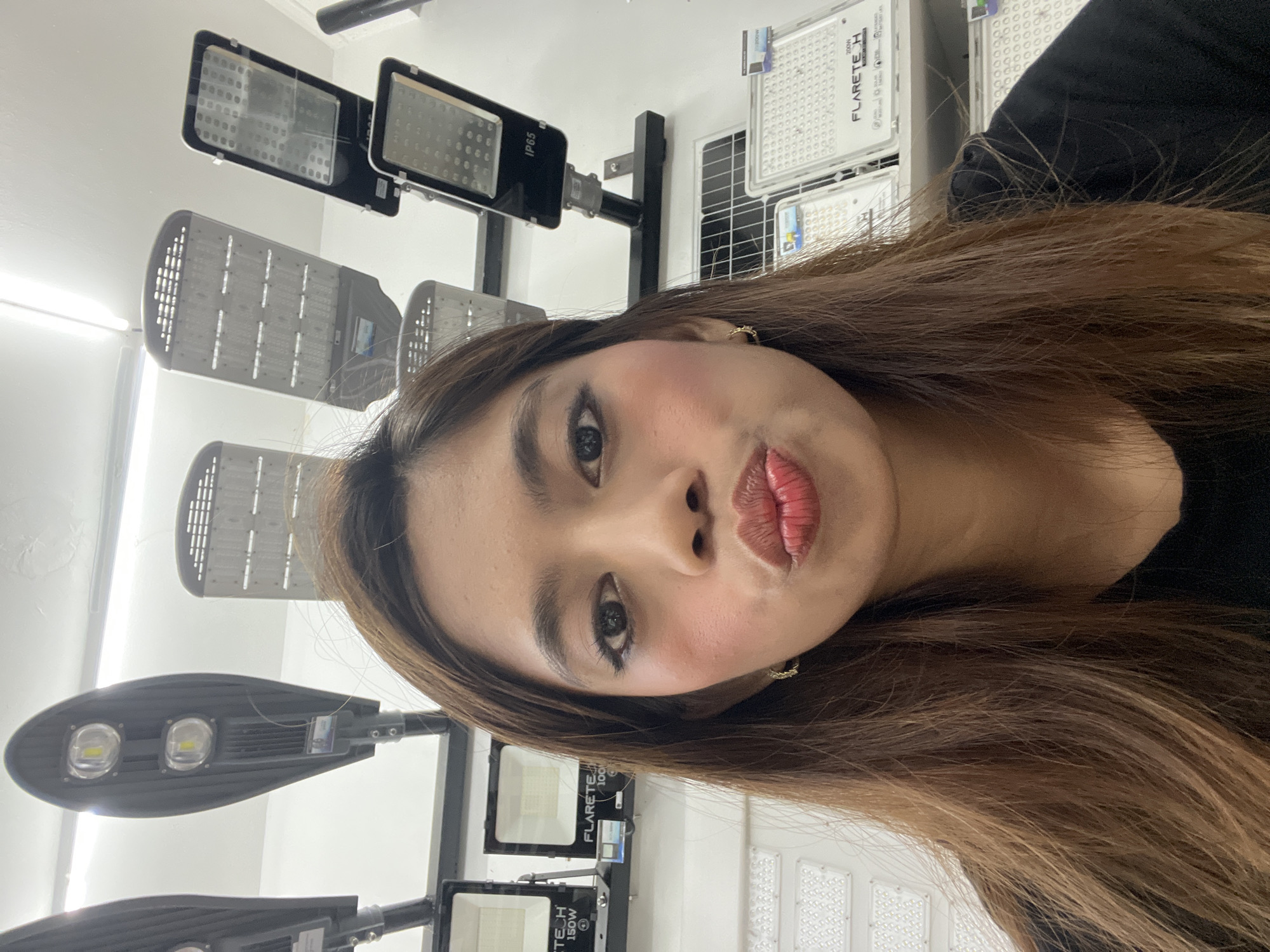 A selfie features a woman with long, brown hair and a light complexion, looking directly at the camera with a "pouty" expression. She has dark eye makeup, rosy blush on her cheeks, and reddish-brown lipstick. A small, gold-colored hoop earring is visible on her right ear. She is wearing a black top or clothing, with her hair falling over her shoulders.
The background reveals an indoor setting, likely a showroom or store in Davao City, Philippines, dedicated to selling outdoor lighting equipment. Several types of light fixtures are displayed on vertical stands against a light-colored wall. These include elongated street lights and rectangular floodlights, many of which appear to be LED-based, indicated by their array of small light-emitting diodes.
Visible text on the products provides further details. One black street light fixture clearly shows "IP65," indicating its ingress protection rating suitable for outdoor use. Several fixtures and their packaging feature the brand name "FLARETE H." Associated with these are wattage indicators such as "200W" and "150W," suggesting varying power outputs. The word "SOLAR" is also visible on one of the "FLARETE H" labels, implying that some of the displayed products are solar-powered lighting solutions. The overall scene suggests a retail environment for modern lighting technology.