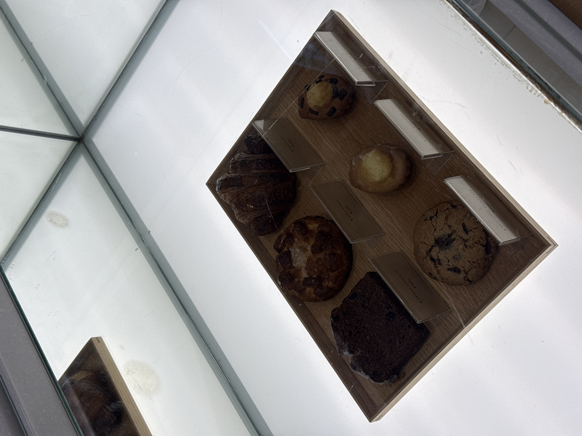 The image shows a display case of baked goods in a bakery, potentially located in Seoul, South Korea, based on the signage accompanying the pastries. The display is a rectangular wooden tray with compartments, each containing a different type of pastry, including a croissant, a chocolate chip cookie, and a brownie. Each pastry has a clear plastic label with pricing and product information, possibly in Korean and English. The display is likely located in a commercial setting, inside a glass case. The lighting is bright, giving the scene a clean and polished appearance. The image is taken at an angle, adding visual interest.