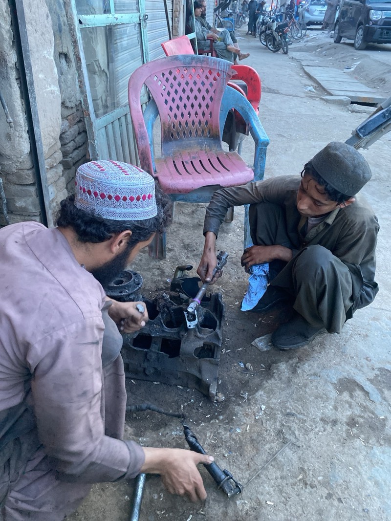 This medium eye-level shot captures a street scene in Kandahar, Afghanistan, focusing on two individuals engaged in repair work on a piece of machinery. In the foreground, two men, or possibly an adult and a youth, are crouched over a dark, heavy engine block or similar mechanical part lying on the dirt and concrete ground. The man on the left, seen from behind, wears a light pinkish-brown loose shirt and trousers, with a prominent white and red patterned kufi (skullcap) on his head. He holds a metal tool in each hand, appearing to be in the process of disassembling or assembling something. Beside him, to the right, a younger individual with a dark cap on his head is also crouched, wearing a dark olive-green loose garment. He is using a wrench-like tool on a component of the engine block, with a piece of blue cloth nearby.
In the midground, behind the workers, a stack of worn plastic chairs – a pink one on top of a blue one, with a red one partially visible behind – stands against a rough, light-colored wall with a greenish metal shutter or door. To the left, part of a crumbling brick structure is visible. Further in the background, a man is seated on one of the red plastic chairs, observing the street.
The scene extends into a dusty street or alleyway where several people are visible, some on motorcycles. Various vehicles, including cars, are parked or moving in the distance. The lighting suggests daytime, possibly late afternoon, with a generally muted and somewhat dusty atmosphere. The clothing, architecture, and overall setting are consistent with the location context of Kandahar, Afghanistan. No discernible text is visible in the image.