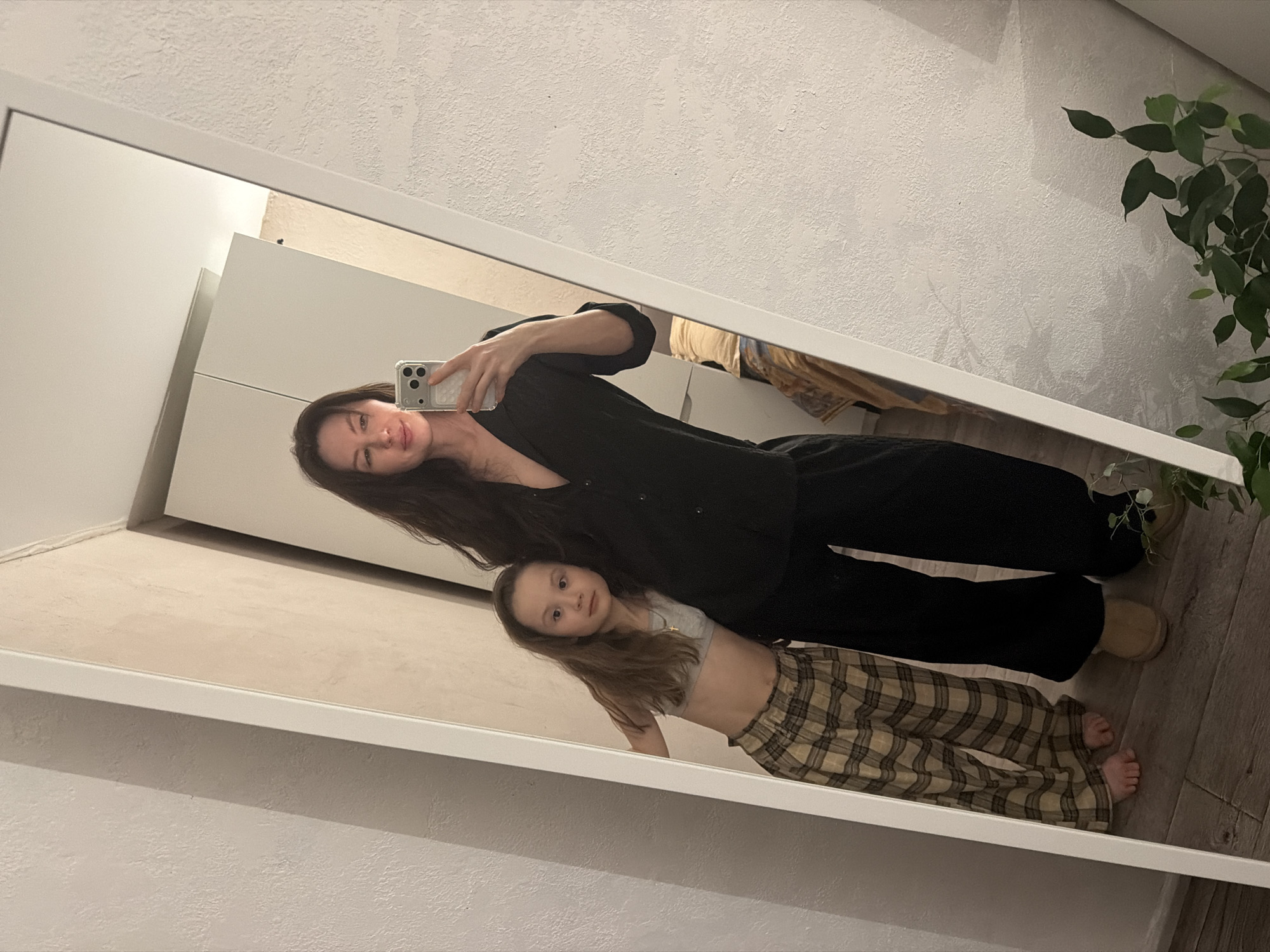 A woman with long dark hair wearing black clothes is taking a selfie with a young girl in a mirror. The girl has long brown hair and is wearing a light-colored top and plaid pants. The setting appears to be indoors, with a white wall and a plant visible in the reflection. There is no explicit information to confirm it is in the city of Perm, Russia.