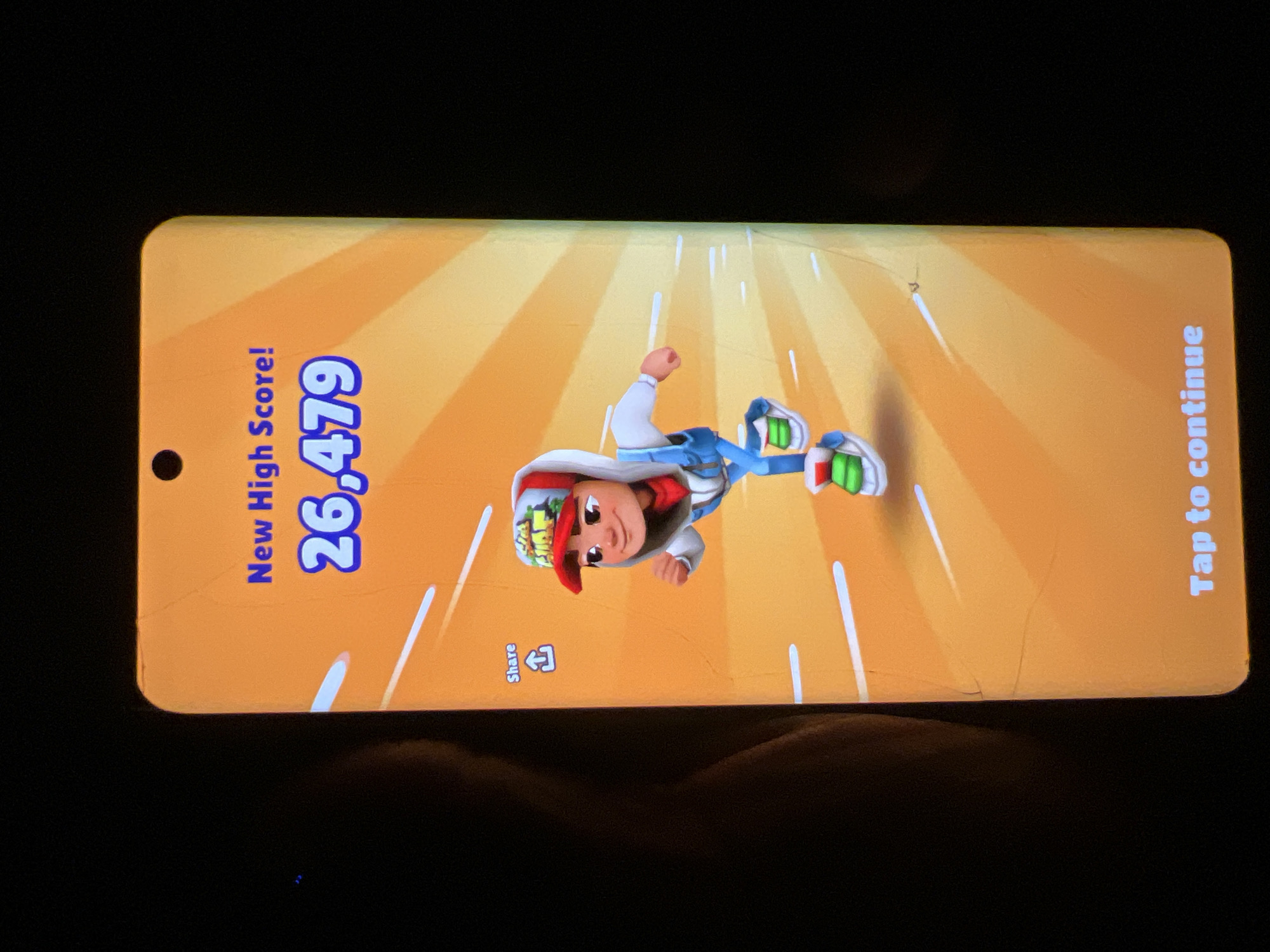 This image shows a mobile phone screen displaying a "New High Score!" message from the game Subway Surfers. The score achieved is 26,479. A character from the game is depicted running. Below the character, there is a "Share" button with an upload icon. At the bottom of the screen, the text "Tap to continue" is visible. The background of the game interface is orange with radiating lines. The phone itself is held in a dark environment, with a dark blurry shape to the left, possibly a person or an object. The screen has some visible cracks and scratches. The location is not discernible from the image provided.