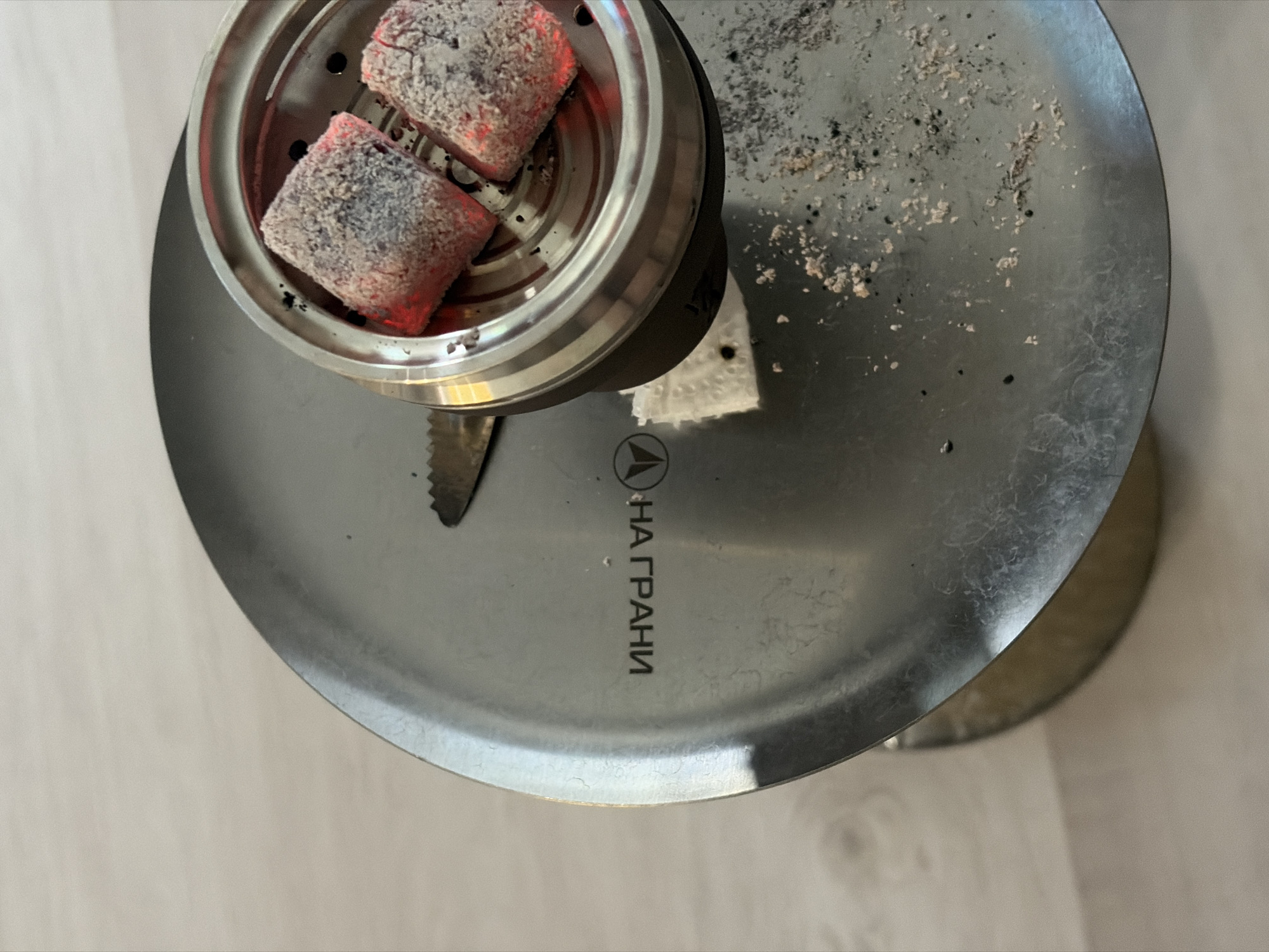 An overhead shot shows a hookah with two glowing red-hot charcoal cubes resting in a metal heat management device. Ash and residue cover the large circular hookah plate. The Russian inscription "НА ГРАНИ" is visible on the plate. The hookah sits on a neutral-colored, possibly wooden, surface. The setting appears to be indoors. There is no explicit evidence linking the scene to Izhevsk, Russia.