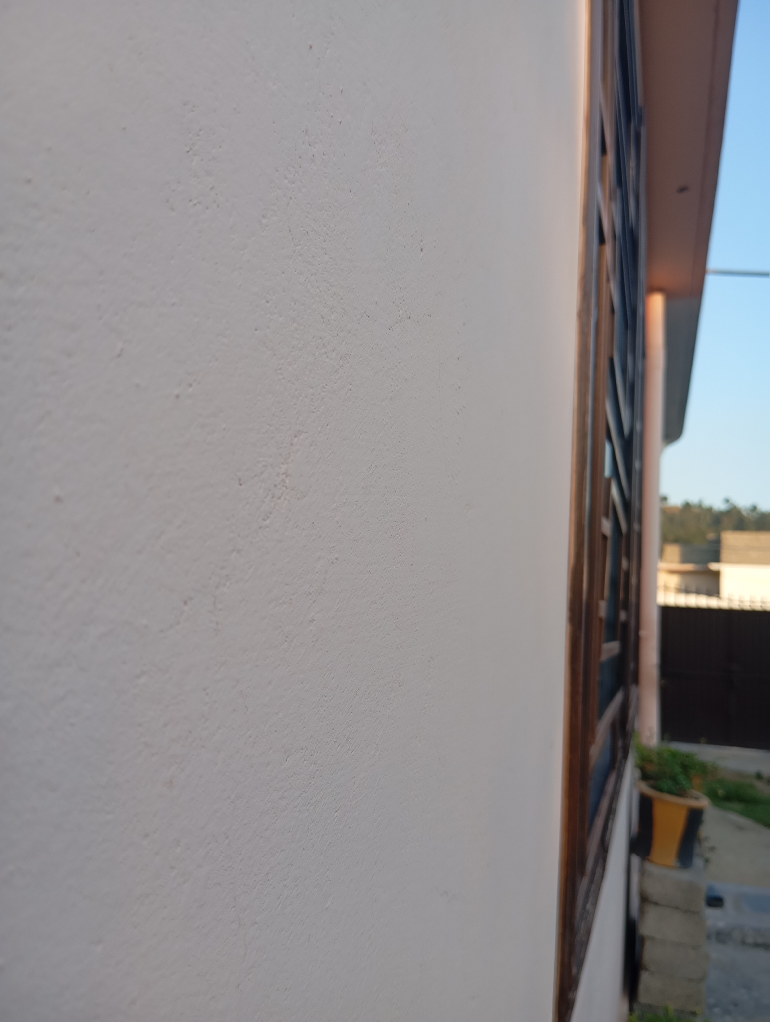 A close-up, side-view of a textured, off-white wall of a building extends from the left to the right edge of the frame, occupying the majority of the image. To the right of the wall, a portion of a window frame with dark, vertical bars is visible. Beyond the window, the building's trim is a light brown, and a light pink overhang is present. The background shows a blurry landscape with green trees and structures under a clear blue sky. In the lower right corner, a yellow and black planter with green plants sits on top of stacked grey bricks.
