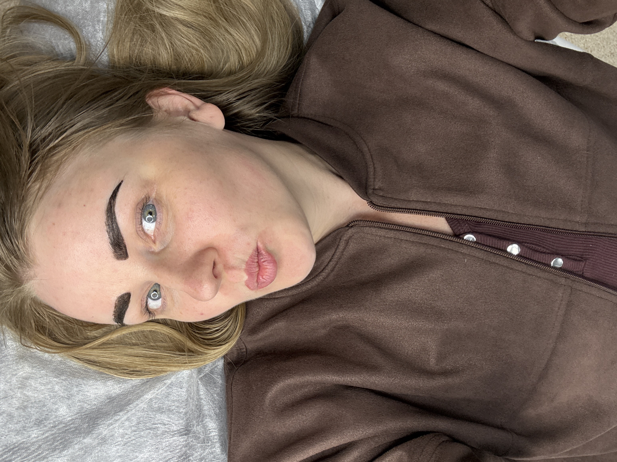 The image shows a woman lying on a gray surface, possibly a medical examination table, wearing a brown zip-up jacket over a burgundy top with silver buttons. She has blonde hair and striking blue eyes. Her eyebrows appear to be freshly tattooed or filled in with dark makeup. The image may have been taken in Chaikovsky, Russia, but this cannot be verified from the image alone. The scene is a close-up shot focused on the woman's face and upper body.