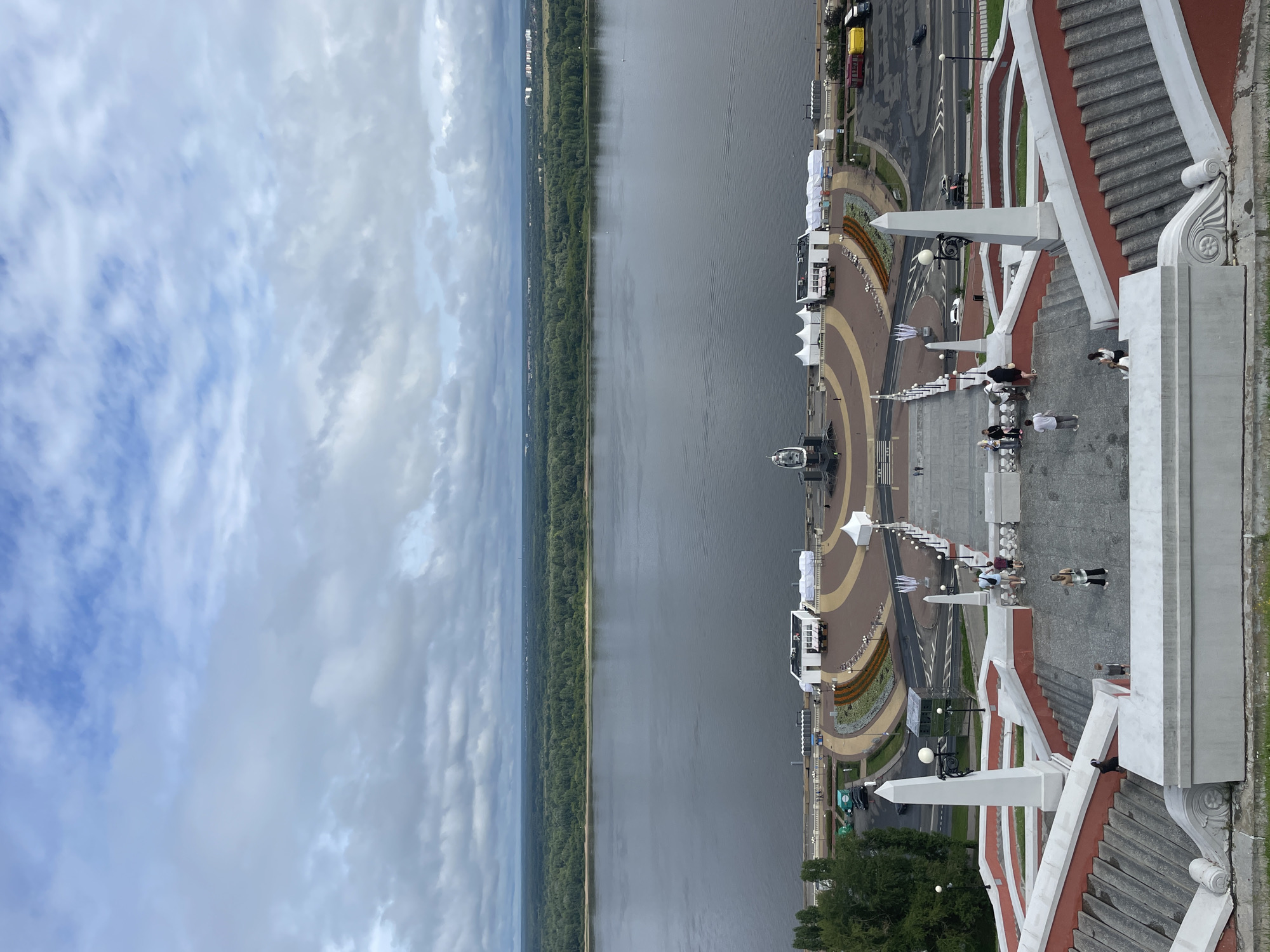 The image is a high-angle, wide shot of the Chkalov Stairs in Nizhny Novgorod, Russia. The stairs are a prominent architectural feature leading down to the Volga River. The foreground shows the upper portion of the wide, multi-tiered stone staircase, with people visible on the steps. The stairs are decorated with red and white trim. Two obelisk-like structures with lamps stand on either side of the stairs.
Below the stairs is a wide, paved plaza with various temporary structures, including white tents and a white building. A monument is visible in the center of the plaza. A road with cars and parking spaces is to the right of the plaza.
The middle ground is dominated by the expansive Volga River, its surface calm with gentle ripples. A densely forested shoreline is visible on the opposite bank of the river. The background consists of a flat, green landscape with scattered buildings and a hazy sky filled with gray and white clouds. The overall scene suggests an overcast day.