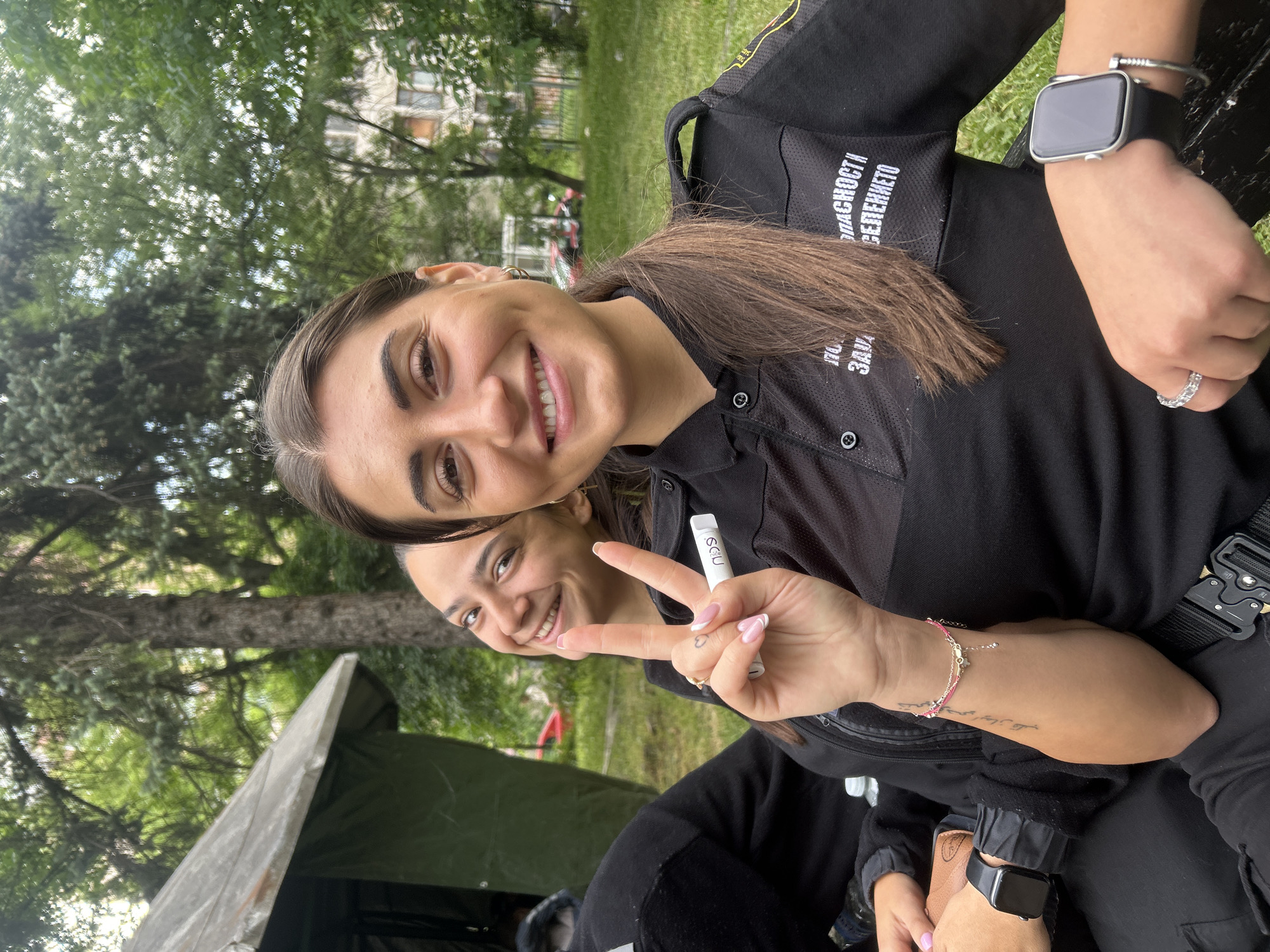 Two young women are posing for a photo outdoors. The primary subject in the foreground is smiling broadly at the camera while holding up a white vaping device and making a peace sign with her fingers. She has dark hair, fair skin, and is wearing a black uniform shirt with lettering in Bulgarian. She also wears a silver Apple Watch and a diamond ring. Behind her, another young woman is visible, smiling slightly. The scene appears to be in a park or green space, with trees and a green tent visible in the background. Based on the uniform and general context, this is likely a volunteer or security event. Given the Bulgarian writing on the shirt, it's likely the location is in Sofia, Bulgaria.