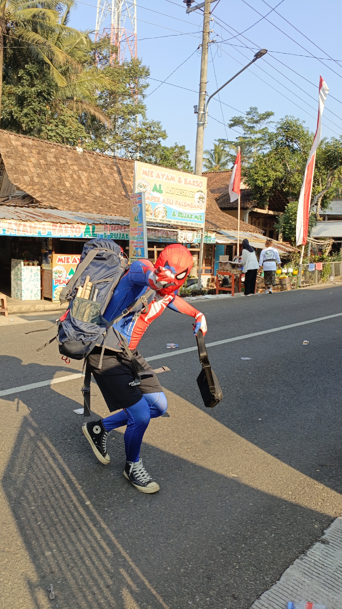 A person in a Spider-Man costume, wearing a large backpack, walks along a paved road in Krajan Lor, Indonesia. The person is wearing black shorts over blue compression pants and black high-top sneakers. They are carrying a black bag in their right hand. The road appears to be a two-lane street with a white line marking the center. On the left side of the road, there is a building with a brown tiled roof and a sign for "MIE AYAM & BAKSO AL AJI LESTARI". There are also palm trees and other vegetation visible. On the right side of the road, there are two people standing near a small structure, and two red and white flags are visible. The sky is clear and blue. The sun is casting shadows on the road.