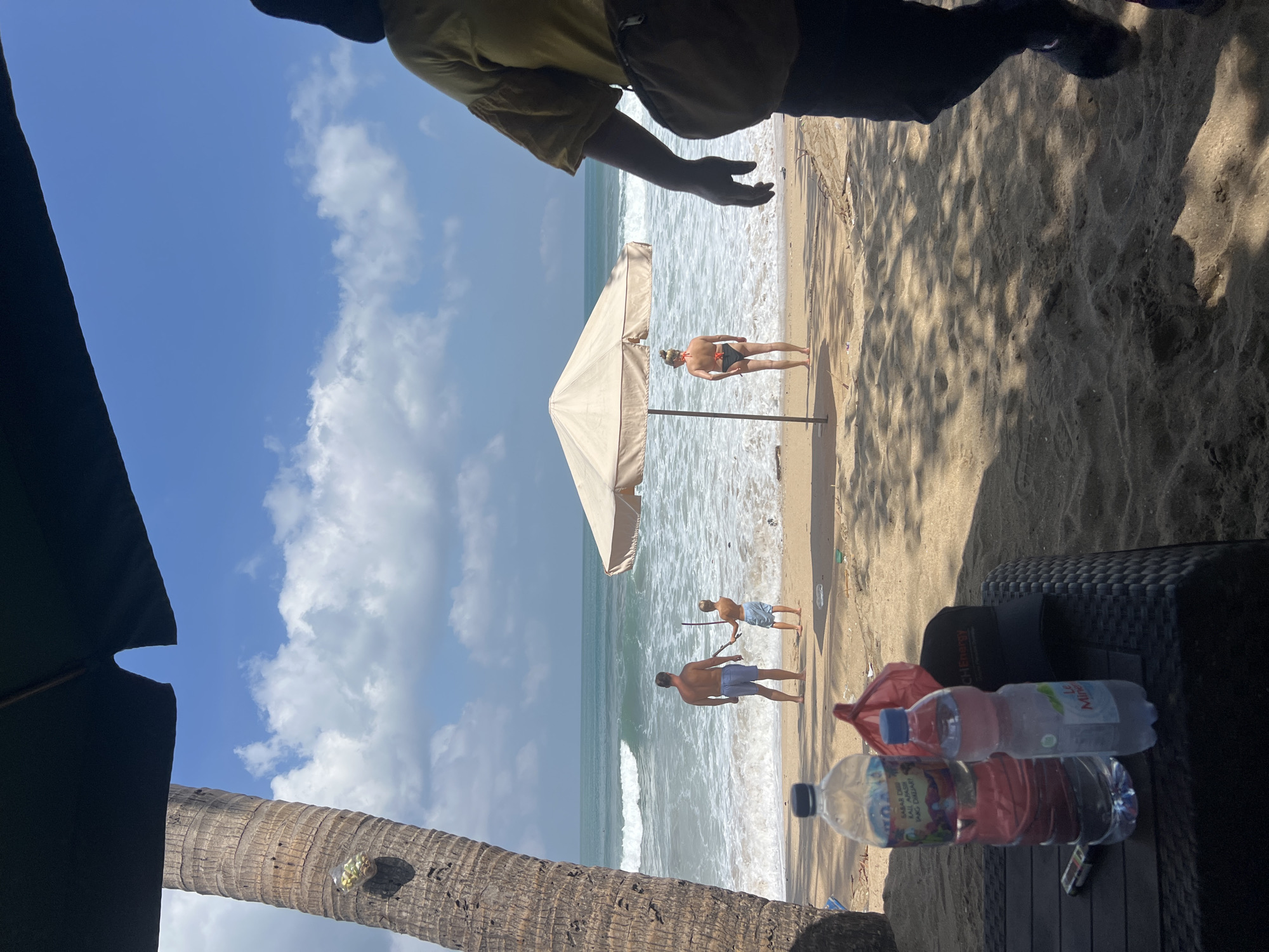 A sunny day at the beach in Kuta, Indonesia, with two men standing near the water and another man and woman near an umbrella. One man is holding a stick. The ocean waves are visible. In the foreground, there are water bottles and a black woven surface. A palm tree trunk is to the left. The sky is blue with some white clouds. The time of day appears to be late morning or early afternoon due to the bright sunlight and shadows. The atmosphere seems relaxed and leisurely. One of the water bottles has "Le Mineral" visible on its label.