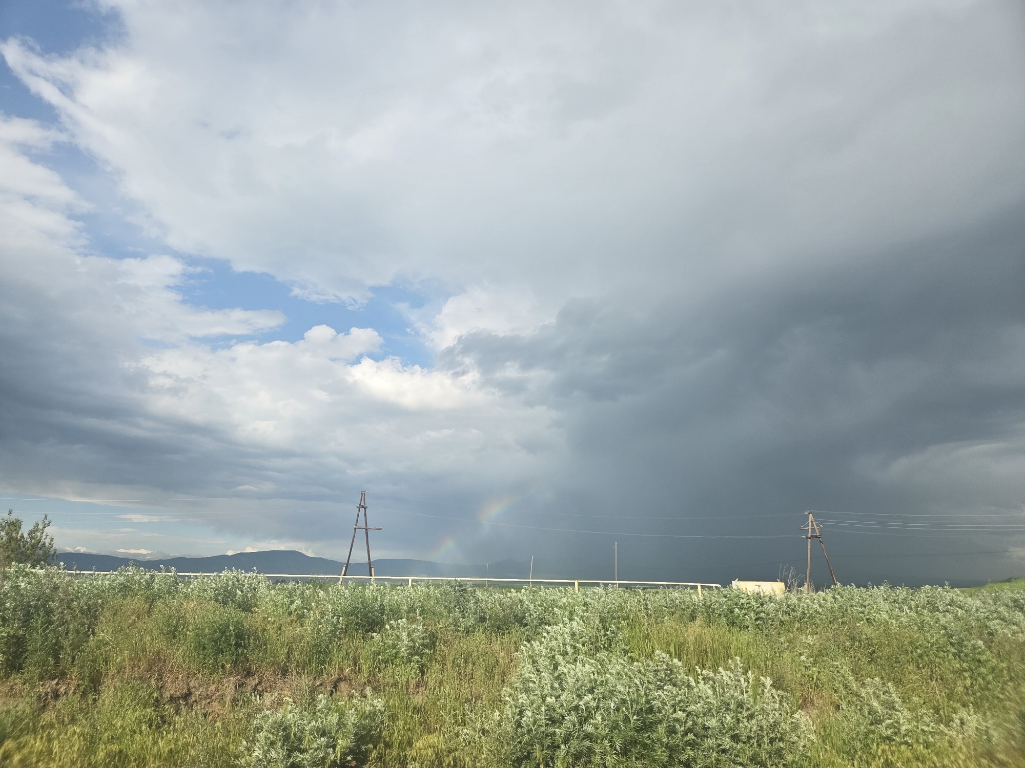This is a landscape photograph taken in the city of Azatan, Armenia. The image features a wide, green field in the foreground, dotted with various types of vegetation and some patches of dry earth. In the mid-ground, there's a white horizontal fence, and behind it, several wooden utility poles are visible, supporting power lines that stretch across the frame. Beyond the fence and poles, dark, ominous storm clouds dominate the sky, contrasting with patches of lighter, brighter clouds and a sliver of blue sky on the left. A faint rainbow is visible in the distance, arching across the stormy sky. In the far background, a range of dark mountains forms the horizon. The scene appears to capture the dramatic atmosphere just before or during a rain shower. There are no people or animals visible in the image.