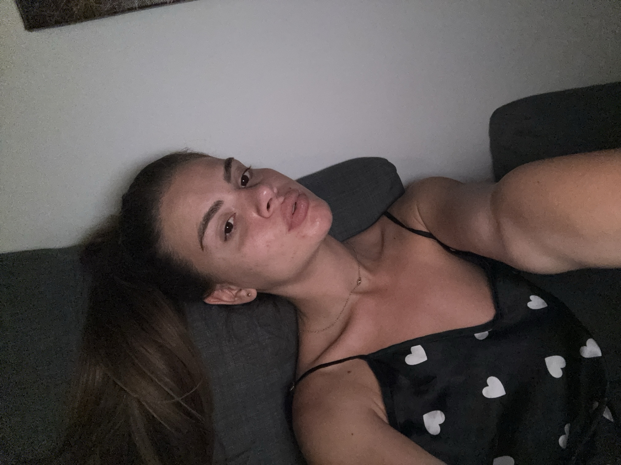 A young woman with brown hair pulled into a ponytail is lying on a dark gray couch. She is wearing a black camisole with white hearts. Her head is tilted back and her eyes are looking towards the camera. The background is a plain, light-colored wall, with a framed picture visible in the top left corner. The scene is indoors and appears to be a residential setting. There is no discernible information within the image to suggest a location in the city of Plovdiv, Bulgaria.