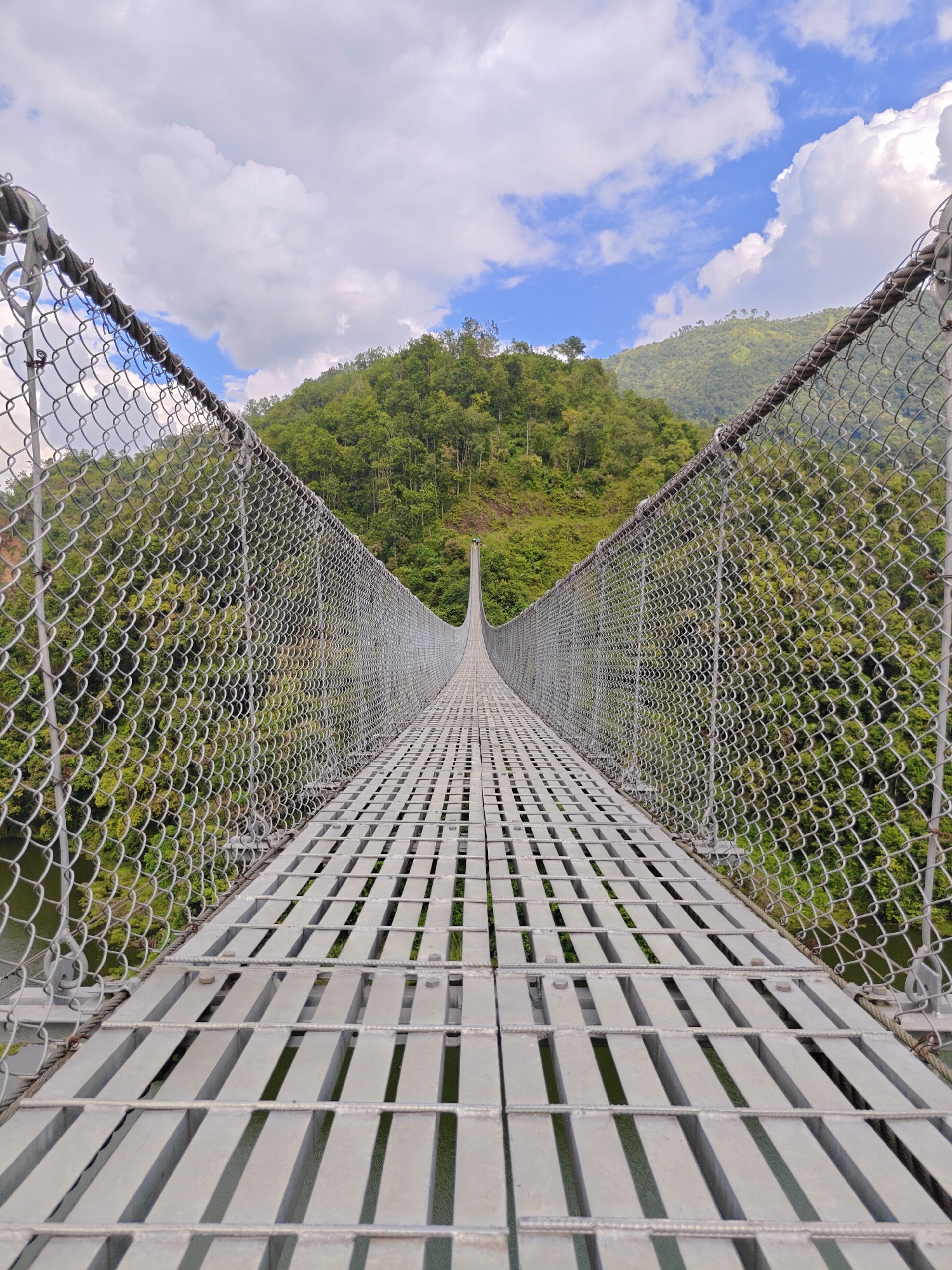 A wide-angle, eye-level shot captures the perspective from the middle of a long, metal suspension bridge in Lekhnath, Nepal. The bridge deck is made of parallel metal slats with gaps between them, revealing a body of water below. Chain-link fencing runs along both sides of the bridge, extending towards the horizon. Beyond the fencing, lush green hills covered in dense trees rise up, meeting a sky with scattered white clouds against a blue backdrop. The perspective of the bridge draws the viewer's eye towards a distant point where the bridge seems to disappear into the landscape. The image is taken during daylight, with natural lighting illuminating the scene. There are no people or animals visible in the frame. The overall scene depicts a natural landscape with human-made infrastructure.