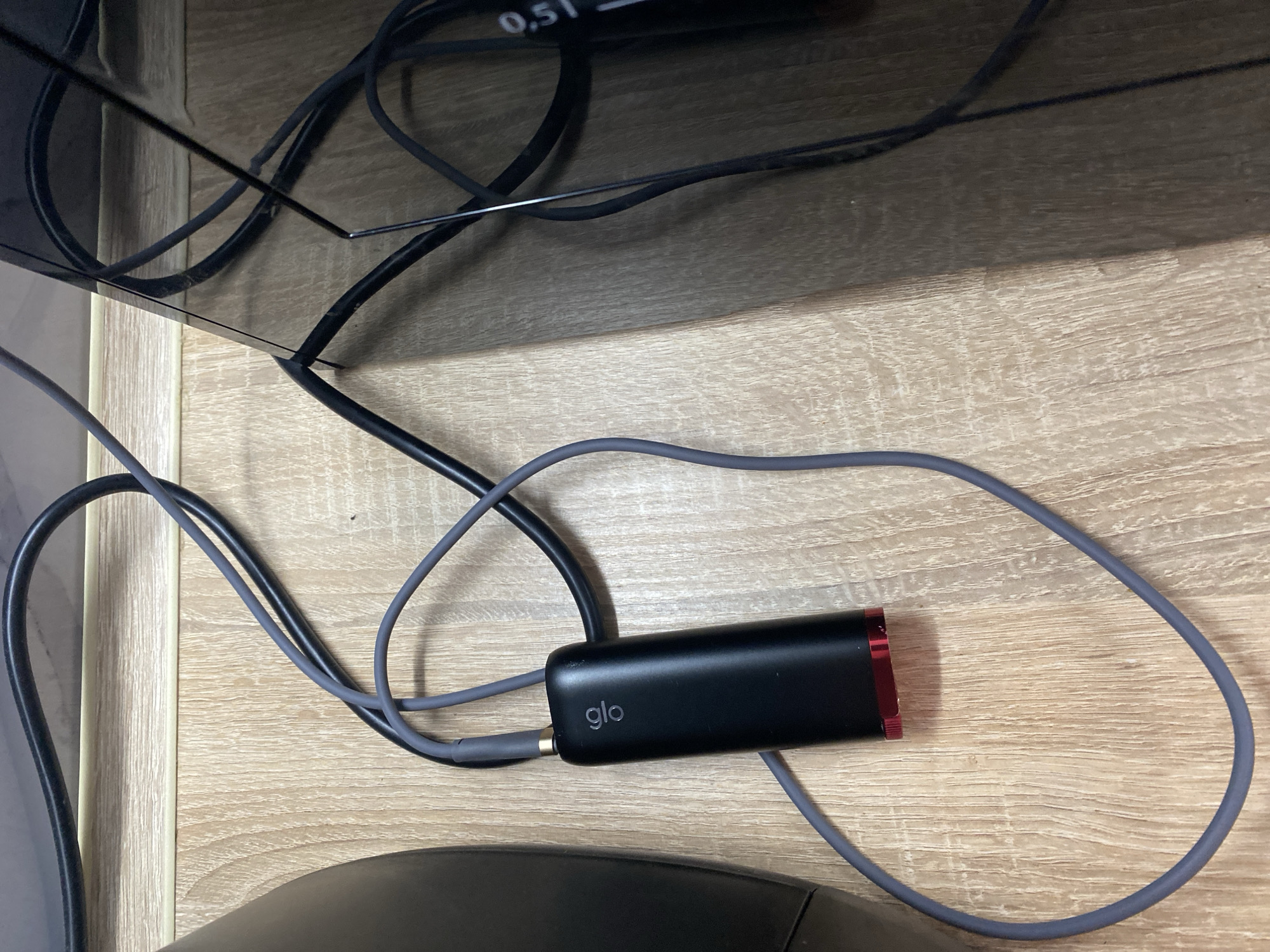 A close-up, top-down shot shows a black Glo device with a red accent at the bottom, connected to a gray charging cable. The device is resting on a light-colored wooden surface, possibly a desk or floor. A black power cable is also visible, snaking across the surface and disappearing behind a dark, reflective object, likely a piece of furniture or electronics. The lighting suggests an indoor environment, with shadows cast across the wooden surface. There is no indication of the city of Kryvyi Rih, Ukraine, or any people present in the image.