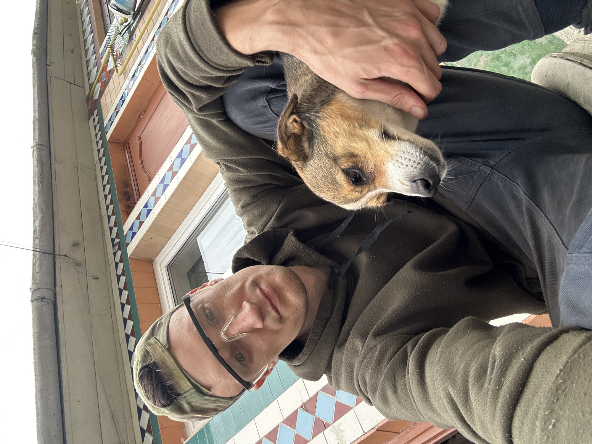 A man wearing a camouflage baseball cap and clear safety glasses is seated outdoors, holding a small dog. The man is wearing a dark green hooded sweatshirt and dark pants. The dog, with tan and white fur, rests its head in the man's lap. They are positioned in front of a building with decorative tiling and a wooden door. The background suggests an urban or suburban setting in Makiv, Ukraine, with a slightly overcast sky.