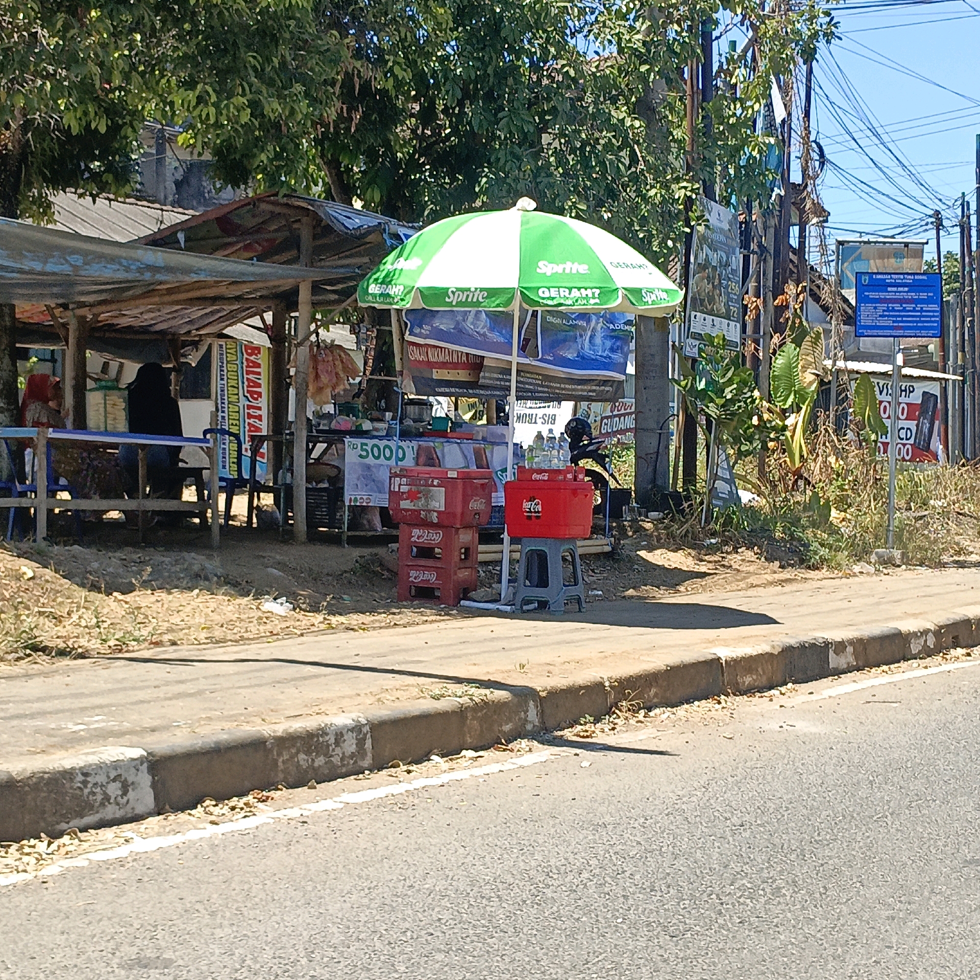 The image depicts a street scene in Salatiga, Indonesia, likely in front of a small roadside stall. The stall features a green and white umbrella advertising "Sprite" and "GERAH?". Below the umbrella, a display of bottled drinks is visible, with some bottles resting on a red Coca-Cola cooler. Adjacent to the cooler, a stack of red Coca-Cola crates is present. In the background, a simple wooden stall structure is visible, with tarps providing shade. Two individuals are partially visible within the stall area; one, wearing a red headscarf, is seated, while another person in black is standing. Various signs are posted on the stall and nearby utility poles, some in Indonesian. The street in the foreground is paved, with a concrete curb and sidewalk separating it from the stall area, which is covered in dry leaves and sparse vegetation. Overhead, a dense network of electrical wires is strung across the sky. The overall scene suggests a hot, sunny day in a commercial area of Salatiga.