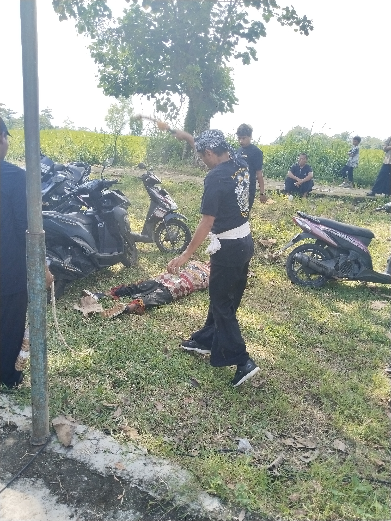 A person is lying on the ground in Mojodoyong, Indonesia, while a man stands over them with a raised hand. The person on the ground is wearing a patterned shirt and pants. The man standing has on a black shirt with white writing, black pants, and a patterned head covering. A white sash is tied around his waist. Several motorcycles are parked nearby. Other people stand or sit in the background, observing. The location appears to be a grassy area near a road or path, with trees and fields visible in the distance. A utility pole is visible on the left.