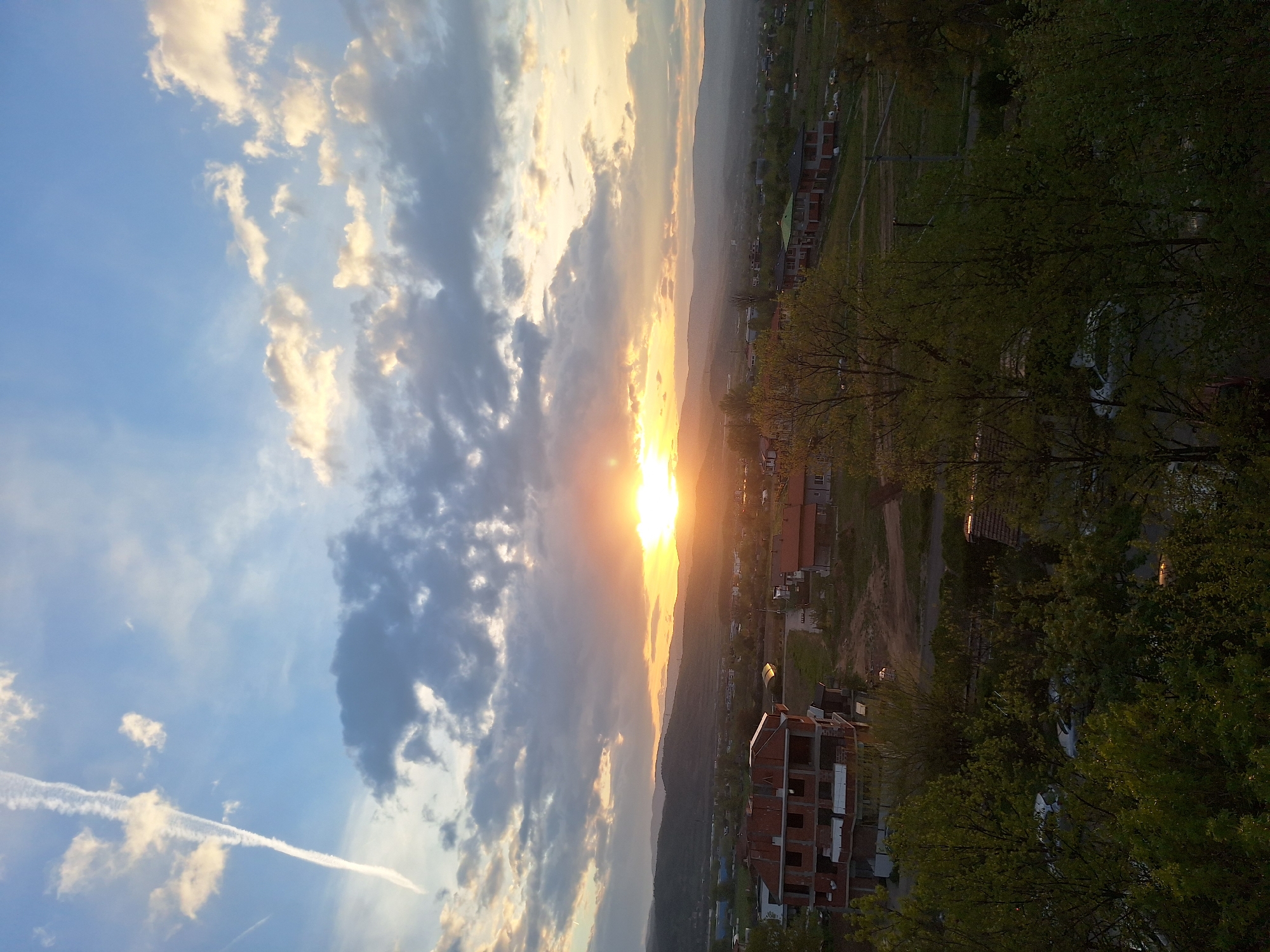This panoramic image captures an outdoor landscape at either sunset or sunrise, overlooking the city of Blagoevgrad, Bulgaria. The scene is viewed from an elevated perspective, showcasing a blend of urban development and natural scenery.
The sky dominates the upper portion of the frame, featuring a gradient from clear blue on the left to warm golden and orange tones near the horizon where the sun is prominently visible. Various clouds are scattered across the sky; some are brightly illuminated by the sun's glow, while others appear darker and more shadowed. A distinct white contrail from an aircraft cuts diagonally across the upper left sky.
Below the horizon, the landscape reveals a valley with distant mountains silhouetted against the bright sky. The midground and foreground are populated with numerous residential buildings and houses. A multi-story building under construction, characterized by exposed brick and concrete framing, is notably visible in the mid-to-foreground on the right. Other completed houses with roofs are also scattered throughout the area. Lush green trees and foliage are abundant, particularly on the right side and in the foreground, indicating a verdant environment, possibly spring or summer. Dirt paths or unpaved roads can be seen winding through the residential areas.
The time of day is clearly either dawn or dusk, as evidenced by the low sun angle and the warm, soft light bathing the scene. The overall atmosphere conveyed is tranquil and picturesque. There are no visible people or discernible activities taking place, and no text is present in the image.