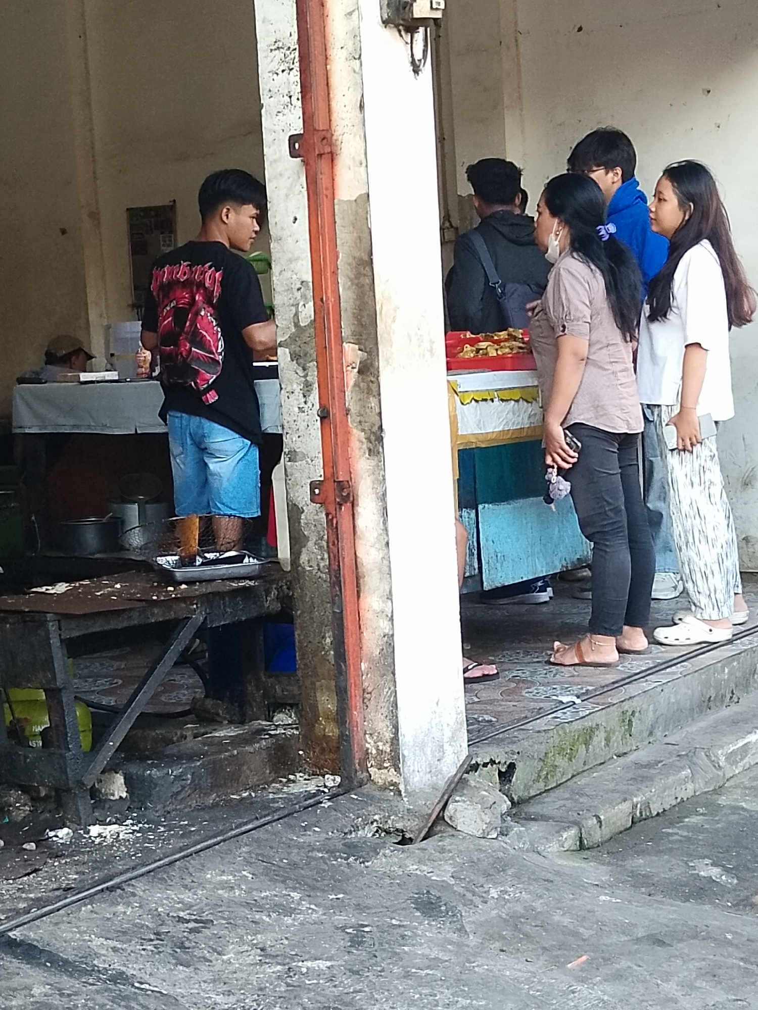 This image captures an outdoor scene in Surabaya, Indonesia, likely a street food stall or local eatery. A young man in a black t-shirt with a graphic print and blue denim shorts stands to the left, facing away from the camera and towards a food preparation area. He is barefoot, with his feet resting on a metal tray containing what appears to be food. Behind him, a person wearing a brown shirt and black pants is seen from the back, with a mask covering their lower face. Next to them, a person in a blue hoodie is looking towards the food. Further to the right, a woman in a white t-shirt and patterned pants stands looking forward. The environment shows a weathered concrete floor with some debris, a metal structure resembling a table or stand with cooking utensils, and a colorful cart with food displayed on it. The overall impression is a busy, everyday scene at a local establishment.