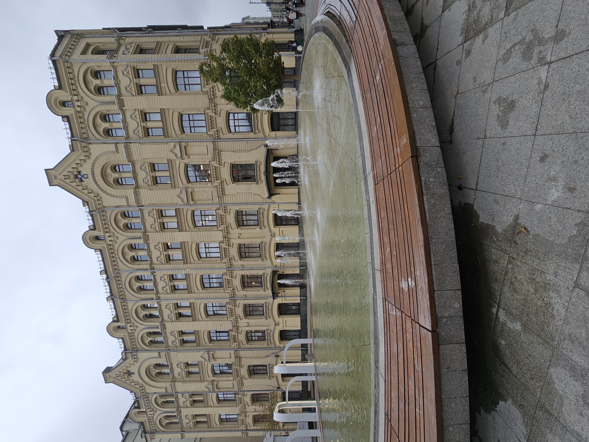 The image depicts a public plaza in Moscow, Russia, featuring a large, circular fountain in the foreground. The fountain has multiple jets of water spraying upwards, creating a dynamic water display. Surrounding the fountain is a curved bench made of wood and stone. In the background, a grand, light yellow building with intricate architectural details stands tall. The building has numerous windows and decorative elements. A lush green tree is visible to the right of the building, partially obscuring it. The ground in the plaza is paved with grey tiles, some of which are wet, suggesting recent rain or fountain spray. The sky is overcast, indicating a cloudy day. The overall scene is one of urban public space, likely a well-maintained area for relaxation and leisure.