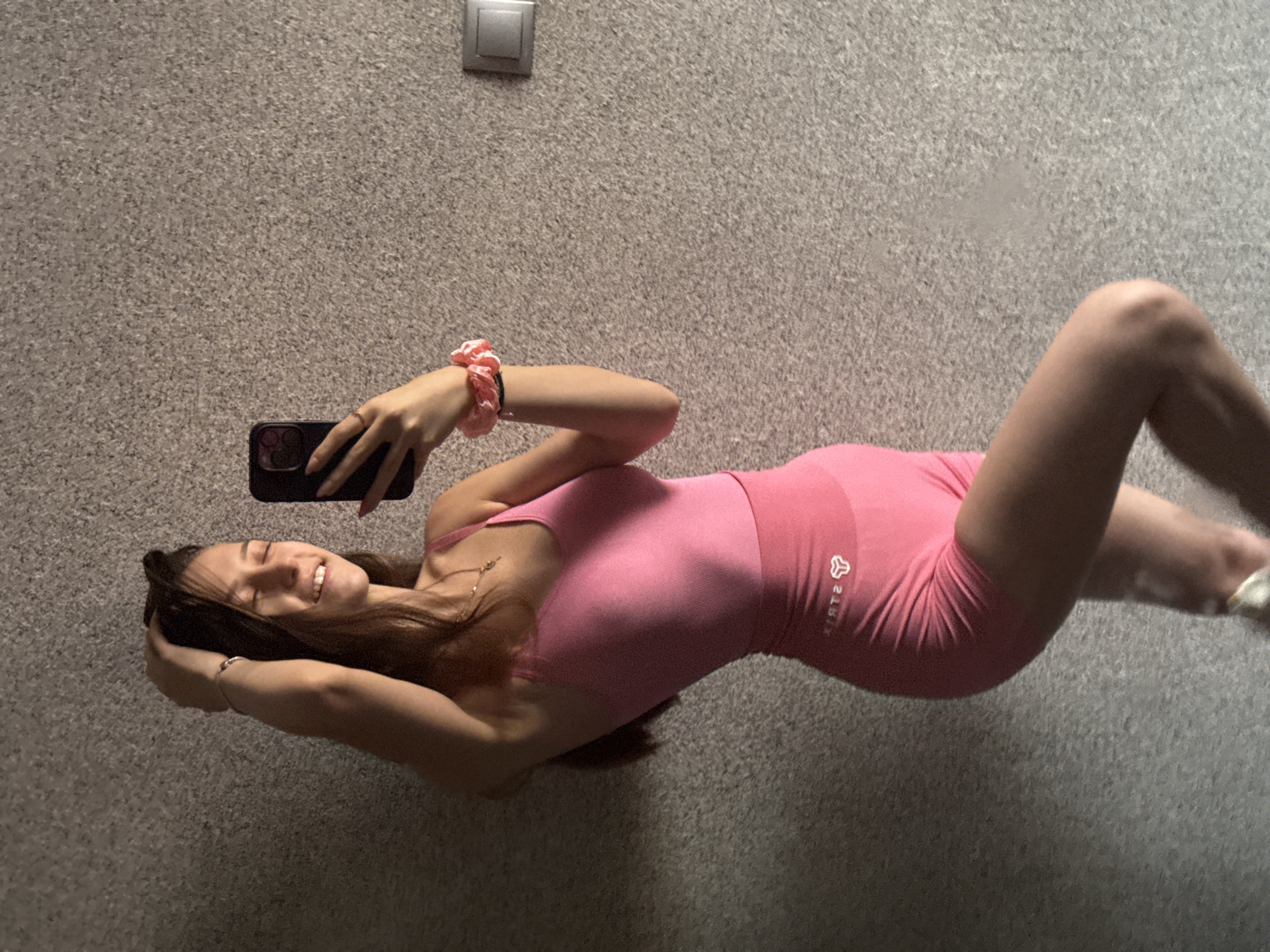 The image shows a young woman taking a selfie in a room with a textured, grey carpeted floor and wall. She is lying down, with her body positioned horizontally across the frame, and her head is angled towards the camera. The woman has long, dark hair, and she is smiling with her eyes closed. She is wearing a pink athletic outfit, consisting of a tank top and shorts. On her wrist, she has a pink scrunchie. She is holding a smartphone in her right hand, using it to capture the reflection in a mirror. A thin, gold necklace is visible around her neck. In the background, a light switch or outlet cover is mounted on the wall. The overall mood of the image is relaxed and casual. The lighting suggests it is indoors, possibly during the day or evening. No specific weather conditions are discernible. No visible text is present in the image. The context provided indicates the location is Sofia, Bulgaria.