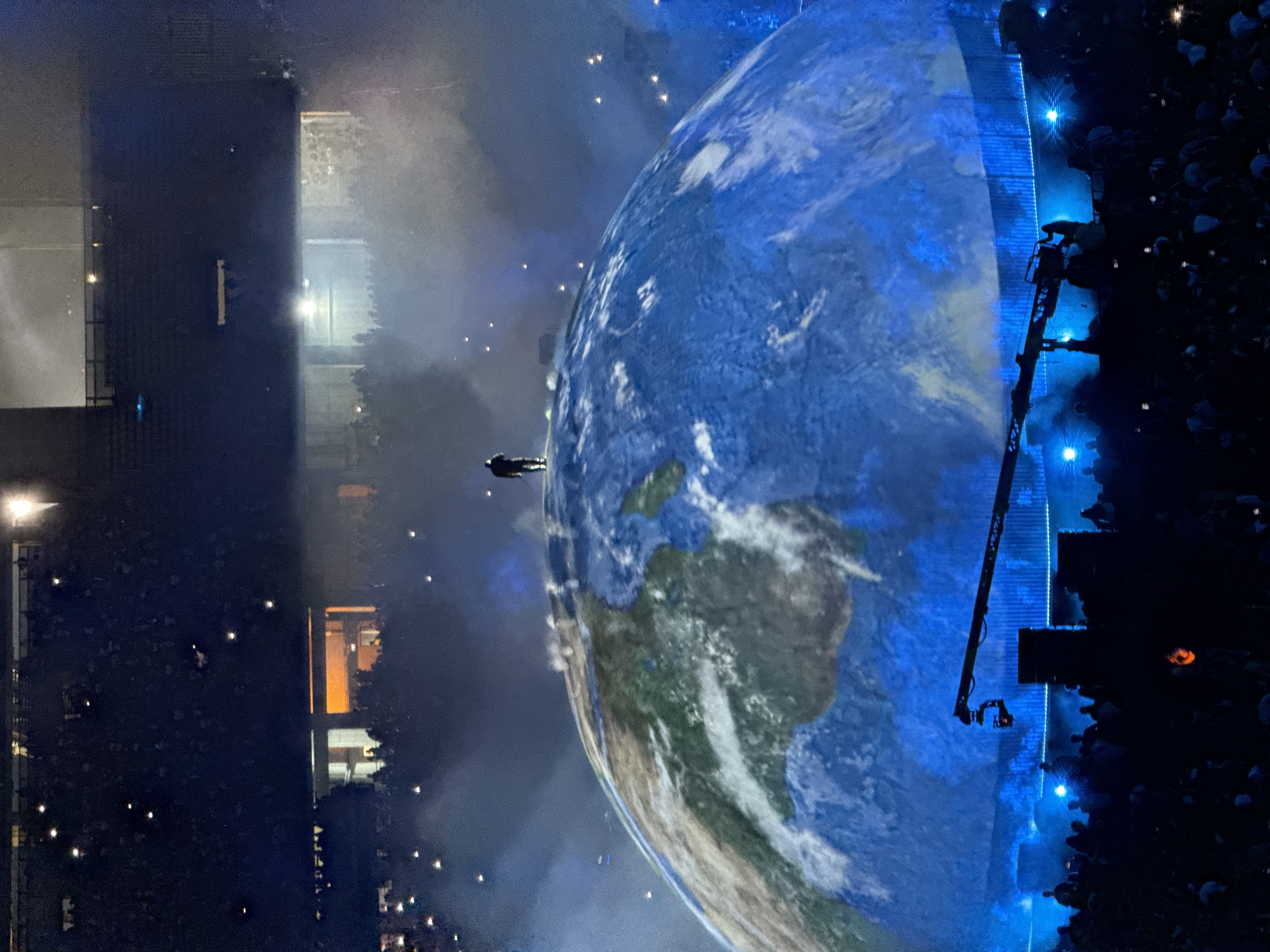 Here is a description of the image:
The image shows a concert or performance taking place in Başakşehir, Turkey. A large screen displays a view of Earth. A performer stands in front of the screen. A crane and camera are positioned on the stage, near a crowd of people. To the left is an apartment complex. The sky is dark, and the scene is illuminated by stage lights and lights from the city and apartment complex.