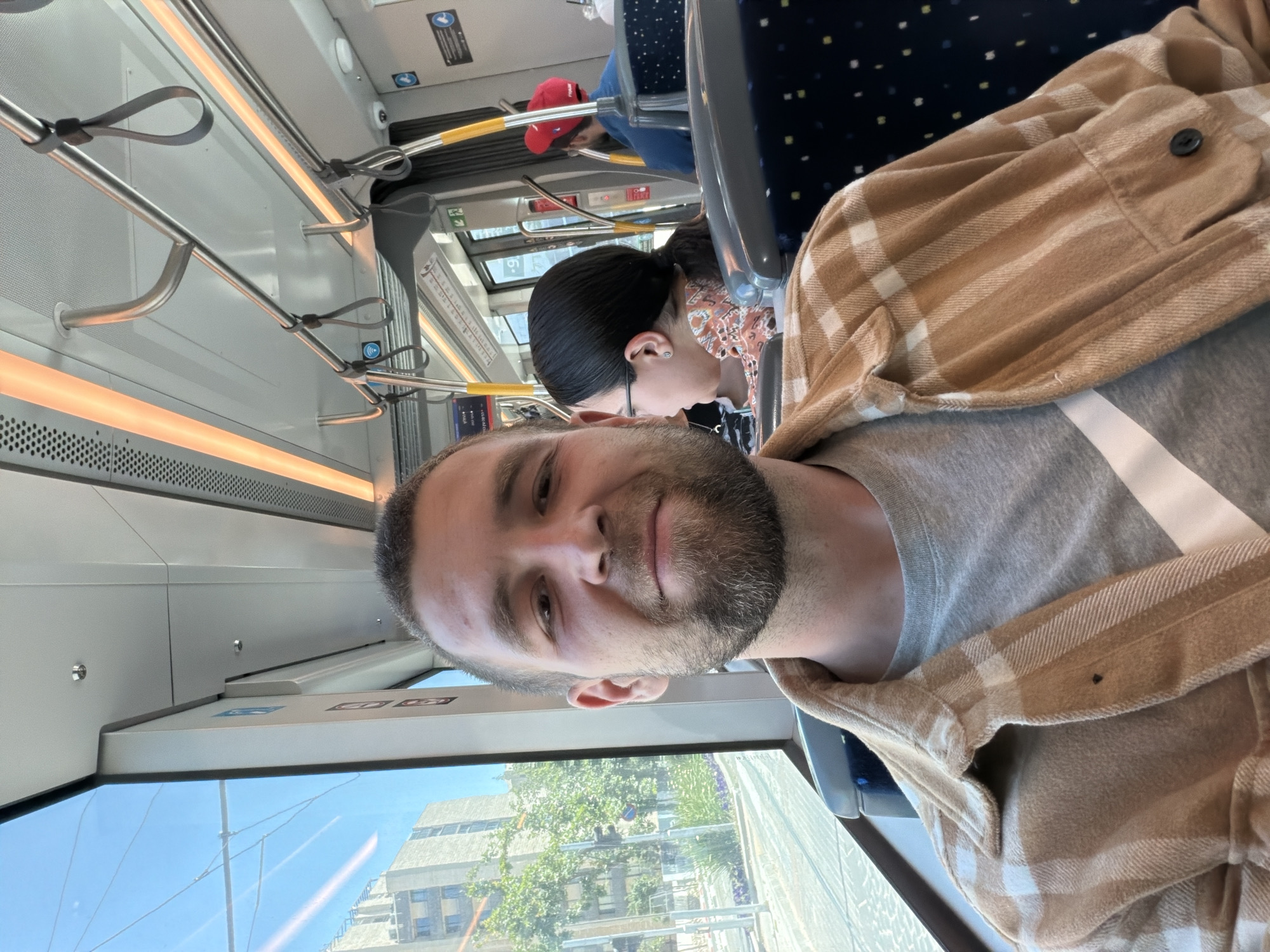 The image depicts the interior of a light rail train traveling in Jerusalem, Israel. A man with a beard is taking a selfie. He is wearing a gray t-shirt and a brown and white plaid shirt. Behind him sits a woman with her hair pulled back in a ponytail. She is wearing a patterned shirt. Further in the background, another passenger wearing a red baseball cap can be seen. Through the train window, buildings and trees are visible. The train's interior features metal handrails and blue and gray seats.