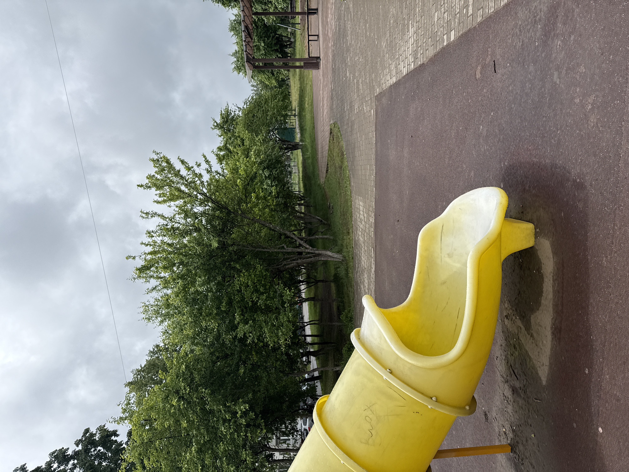 The image depicts an outdoor playground or park scene in Chaikovsky, Russia, under an overcast sky. In the foreground, a bright yellow children's slide, appearing to be the exit of a tunnel slide, rests on a dark reddish-brown, textured ground, likely a rubber safety surface. Faint white graffiti, spelling "Xeny," is visible on the side of the yellow slide. Adjacent to the safety surface is a light grey paved pathway constructed from bricks or pavers.
Beyond the paved area, a strip of green grass leads to a dense line of trees with vibrant green foliage, forming the background. Partially visible among the trees in the mid-ground is a wooden pergola or shelter structure. The sky is completely covered by grey clouds, indicating an overcast day with soft, diffused lighting. A thin, dark wire stretches across the upper left portion of the sky. No individuals are present in the scene, conveying a quiet and peaceful atmosphere. The abundant green leaves suggest the season is likely late spring or summer.