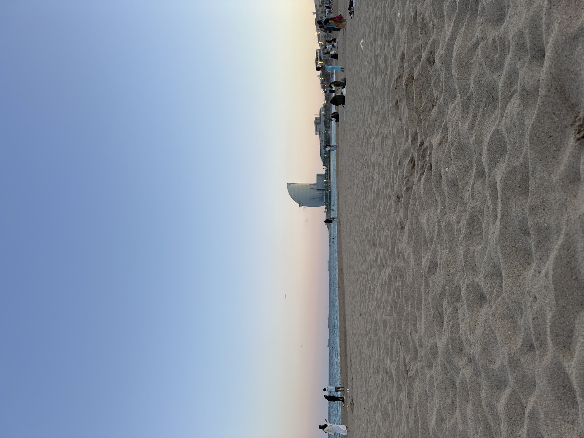 The image shows a beach scene in Barcelona, Spain, at sunset. The sky fades from light blue to pink and orange near the horizon. The beach extends from the bottom right to the middle of the image, covered in textured sand. A building with a sail-like structure is visible in the distance along the shoreline. The Mediterranean Sea appears as a thin strip between the beach and the building. People are scattered along the beach, some standing near the water and others further away towards buildings and the city in the background. The image orientation is rotated 90 degrees counterclockwise.