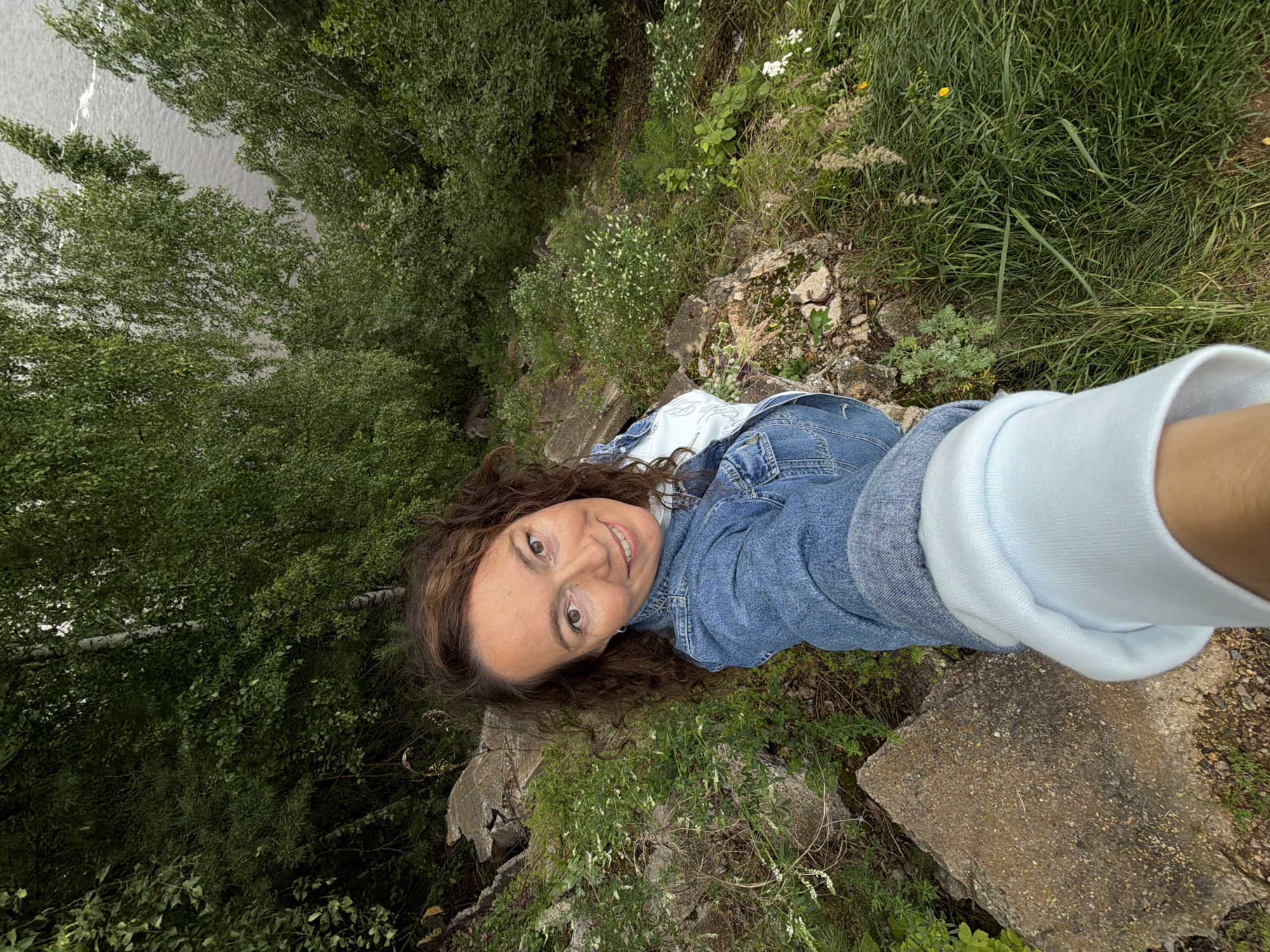 In a selfie taken from a high vantage point, a woman with curly brown hair and light brown eyes smiles at the camera. She is wearing a denim jacket over a light blue hooded sweatshirt. She is positioned on a rocky, overgrown embankment, with large grey stones and patches of green grass and small white flowers. In the background, a dense canopy of green trees, some with white bark, frames a body of water, likely the Kama River, which flows through the city of Chernushka, Russia. The overall scene suggests an outdoor setting in a natural environment near the river.