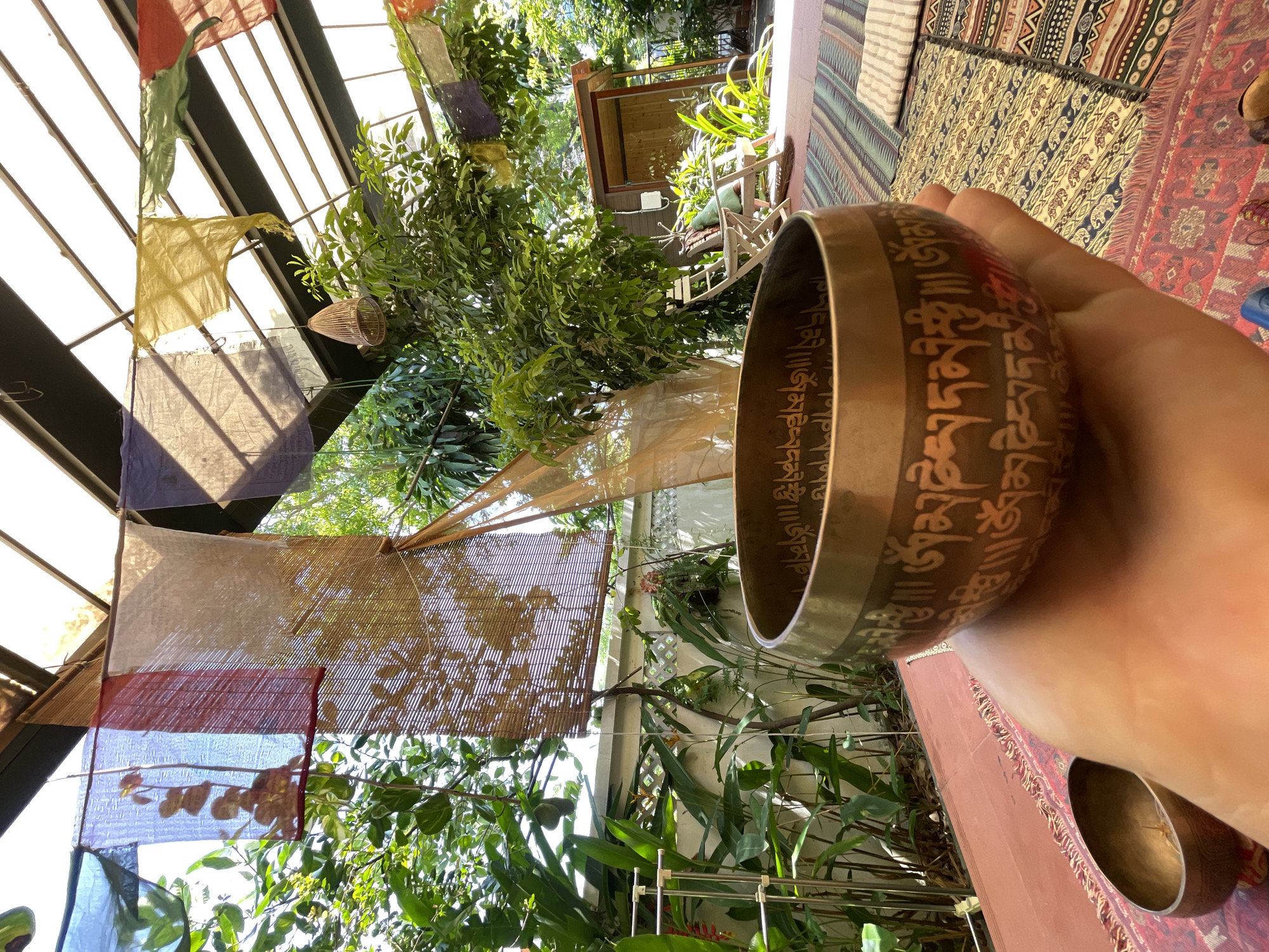 The image captures a close-up of a hand holding a brass singing bowl, adorned with intricate Tibetan script, in what appears to be an outdoor or semi-outdoor seating area. The background suggests a lush, green environment with numerous plants and trees. Prayer flags in various colors are strung across the top of the scene, indicating a possible spiritual or meditation space. In the distance, a small wooden structure and chairs are visible, hinting at a tranquil, possibly residential or retreat setting. The flooring is covered with colorful rugs. The specific location in Đà Nẵng, Vietnam, is not identifiable from the visual information present in the image.