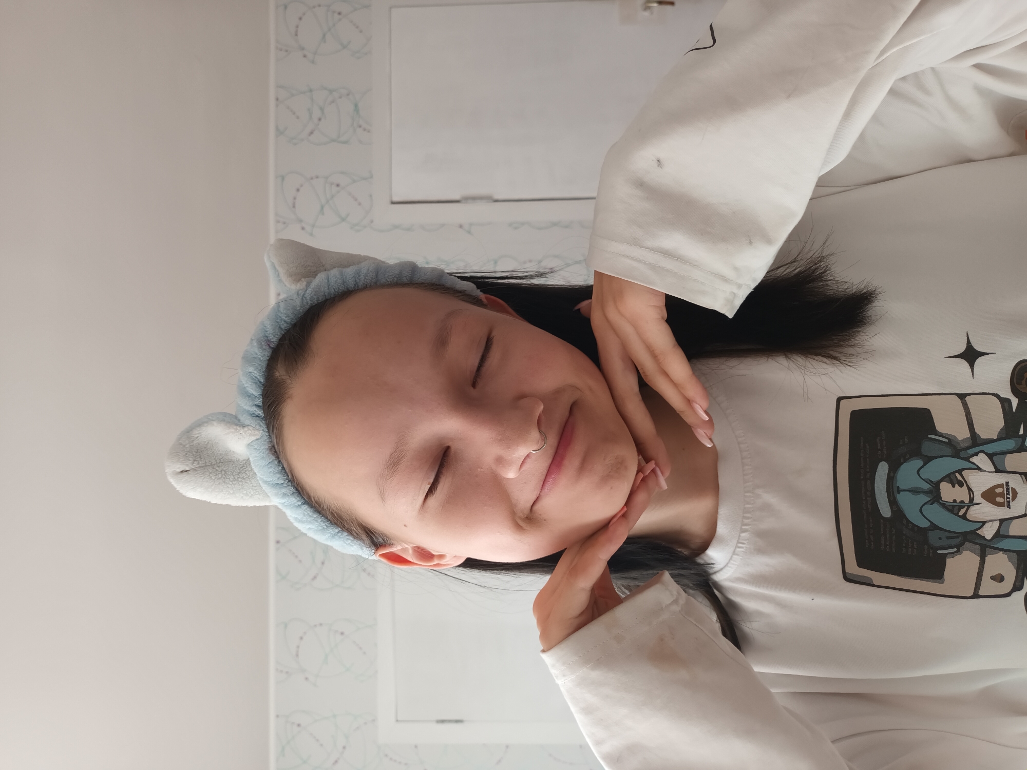A young woman with dark hair and fair skin is posed with her eyes closed, wearing a light blue cat-ear headband. She has a nose ring. Her hands are positioned gently on either side of her face. She is wearing a white, long-sleeved shirt with a cartoon graphic on the front. The background is a wall with a pale blue and white pattern and a white cabinet or door. There is no evidence to indicate the location is in Ольховка, Russia.