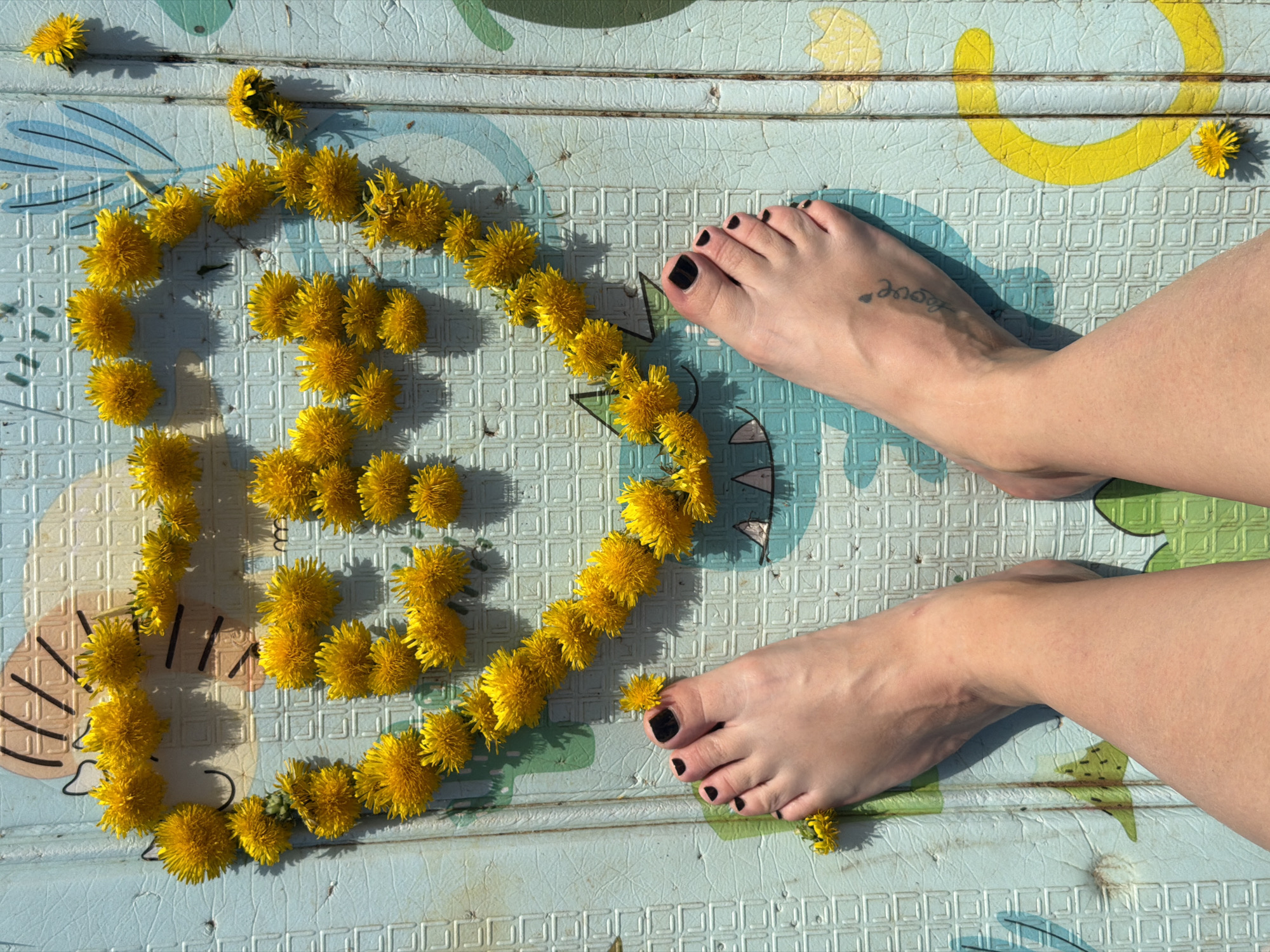 The scene is an outdoor setting, likely a patio or deck, in daylight. The surface is a light blue and white patterned mat with a textured grid pattern, possibly a children's play mat. A string of yellow dandelions, arranged in a rough heart shape, lies on the mat. Two bare feet, toes painted with dark nail polish, are positioned near the top right of the frame. A tattoo is visible on the top of the left foot. A leg extends from each foot, disappearing out of frame on the right side of the image. Additional dandelions are scattered around the frame. The location in the city of Chaikovsky, Russia, cannot be determined from this image.
