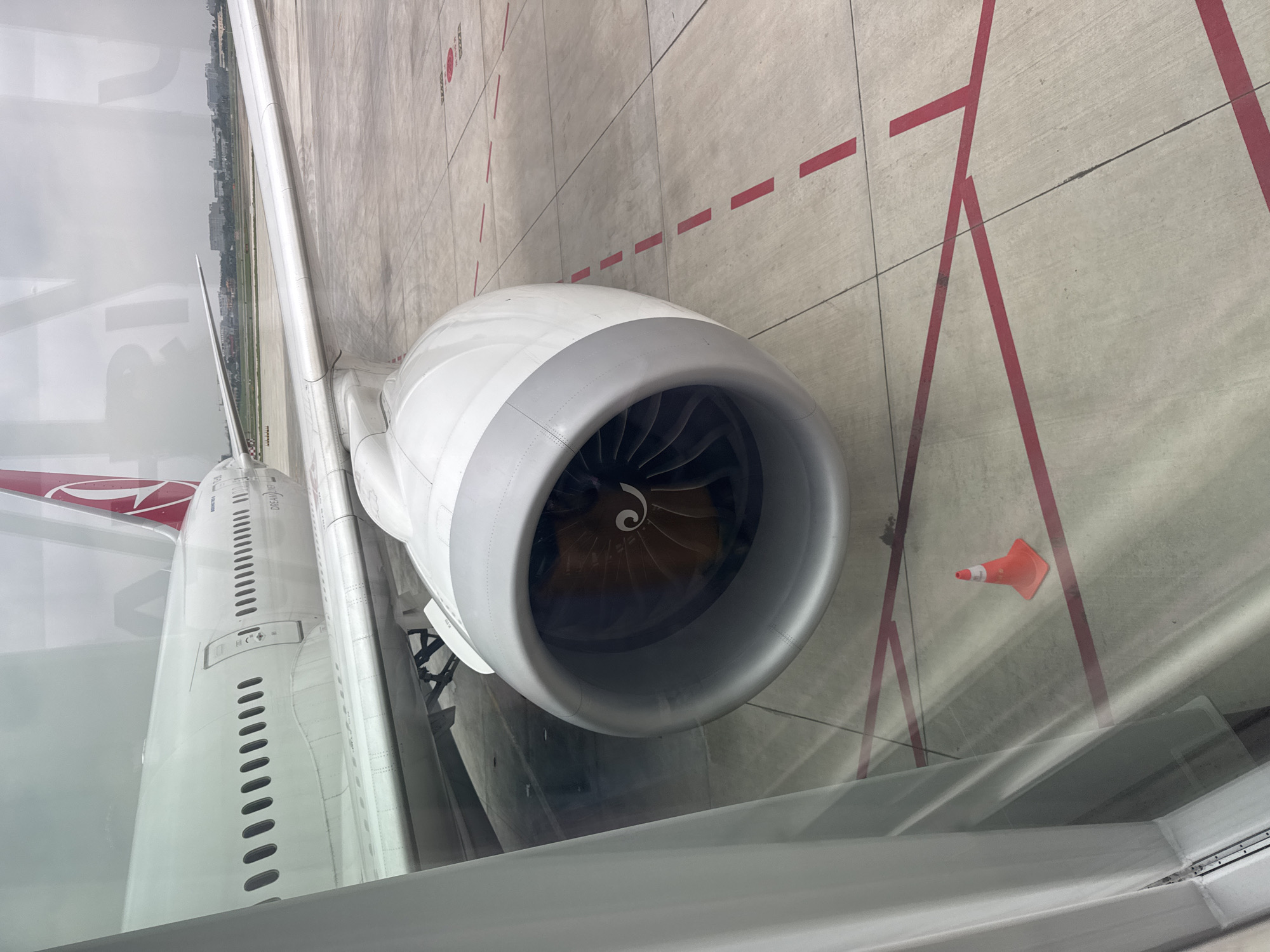 The image is taken from inside an airplane, looking out the window. The focal point is the engine of the aircraft, a large, white and gray structure with a visible turbine inside. To the left, the fuselage of the plane is partially visible, along with a red and white tail. Outside the window, the tarmac is gray concrete with red painted lines. An orange traffic cone sits on the tarmac. The context suggests this is an airport environment, but there is no clear indication of it being in Baiyun District, China, so that cannot be confirmed.