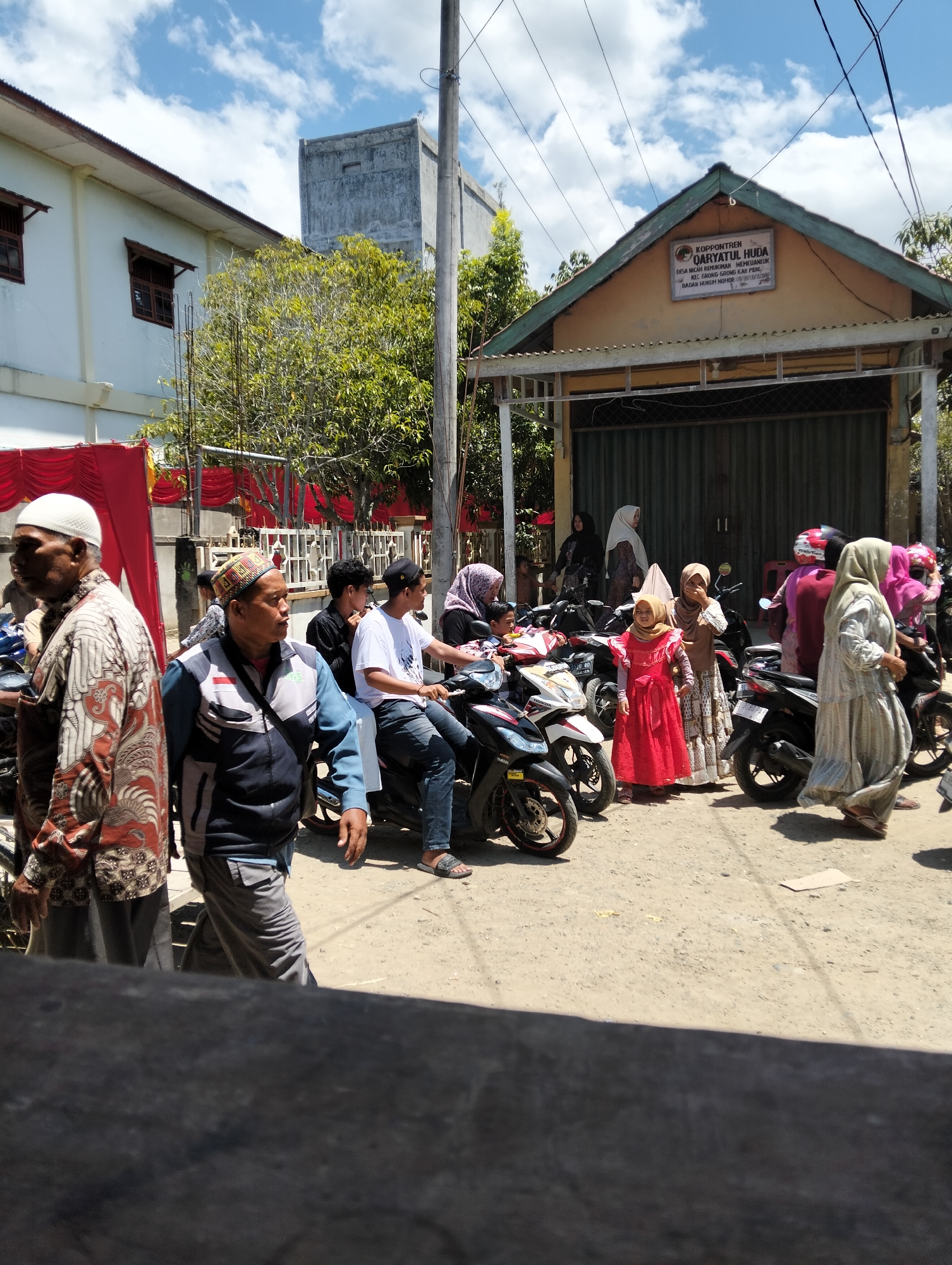 In the city of Teungoh, Indonesia, on a bright, sunny day, a group of people are gathered outdoors. The scene appears to be in front of a building with a sign that reads "KOPONTREN QARYATUL HUDA." Several motorbikes are parked in the foreground and midground, indicating a mode of transportation common in the area.
The people in the image are dressed in various attire, with many women wearing hijabs. Some individuals are standing, while others are seated on the motorbikes. The overall atmosphere suggests a community gathering or event. The background shows a building with a distinctive architectural style, and a tall, concrete structure can be seen in the distance. Lush green trees add to the natural environment surrounding the urban setting. The ground is covered with a dirt or gravel surface, typical of many roads and outdoor spaces in Indonesia. The image captures a moment in the daily life of Teungoh, highlighting its vibrant community and cultural landscape.