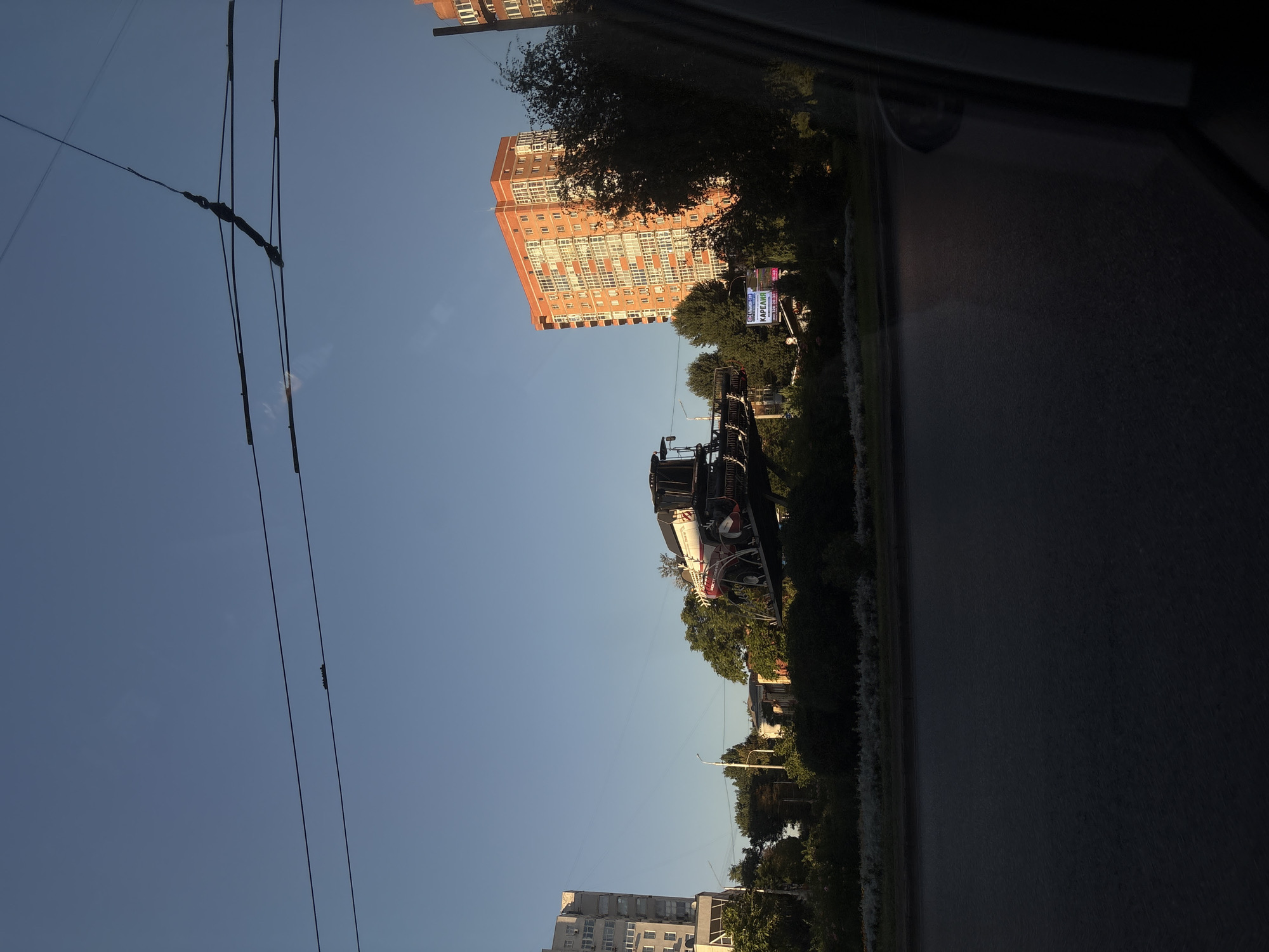 This is a ground-level, medium-shot, eye-level photograph taken from a vehicle, likely a car, looking slightly upwards and towards the right. The scene depicts an urban environment with a clear, blue sky. In the foreground, the dark asphalt of a road occupies the bottom portion of the frame.
Dominating the middle ground and slightly to the left is a sculpture of a combine harvester, mounted on a raised platform. The combine is predominantly white and red, with visible wheels and harvesting mechanisms. Behind the combine, there is a line of green trees and bushes, forming a landscaped area.
To the left of the combine sculpture, a tall, light-colored apartment building with visible balconies and air conditioning units is present. Further to the right, a multi-story residential building with an orange facade and numerous windows stands out. A dark-colored tree with dense foliage partially obscures this building.
Overhead, several black power or trolleybus lines are strung across the sky, crisscrossing the frame. A street lamp is visible behind the combine sculpture. A small billboard with Russian text and an image of a woman is positioned in front of the trees, to the right of the combine. The overall lighting suggests it is late afternoon or early evening, with the sun casting long shadows and highlighting the buildings. Based on the architecture and the presence of a combine harvester as a sculpture, the location is likely Rostov-on-Don, Russia, possibly a monument to agricultural machinery. No people are visible in the frame.