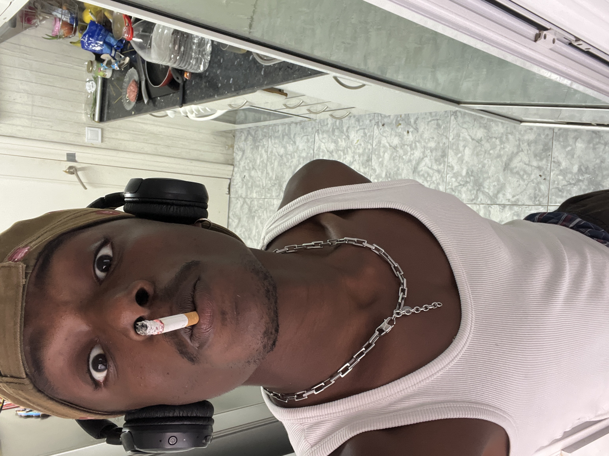 A young Black man with dark skin is prominently featured in a close-up, chest-up portrait, looking directly at the viewer. He wears a white ribbed tank top, a silver chain necklace with rectangular links, and a brown baseball cap turned backward. Black over-ear headphones rest around his neck, with a power button and status light visible on the right earcup. A lit cigarette is held between his lips, emitting a small wisp of smoke. He has short facial hair.
The setting appears to be a kitchen or utility area. The background features light grey mottled wall tiles on the lower half. Above this, white kitchen cabinets with silver handles are visible. A black countertop holds various items, including clear plastic bottles, a pan, and other containers, some containing food items. A white light switch is visible on the tiled wall. A reflective surface or window runs along the top of the frame.
The man is posing for the camera, actively smoking the cigarette. The scene is illuminated by indoor lighting. No visible text is present in the image. The location context for the image is Barcelona, Spain.