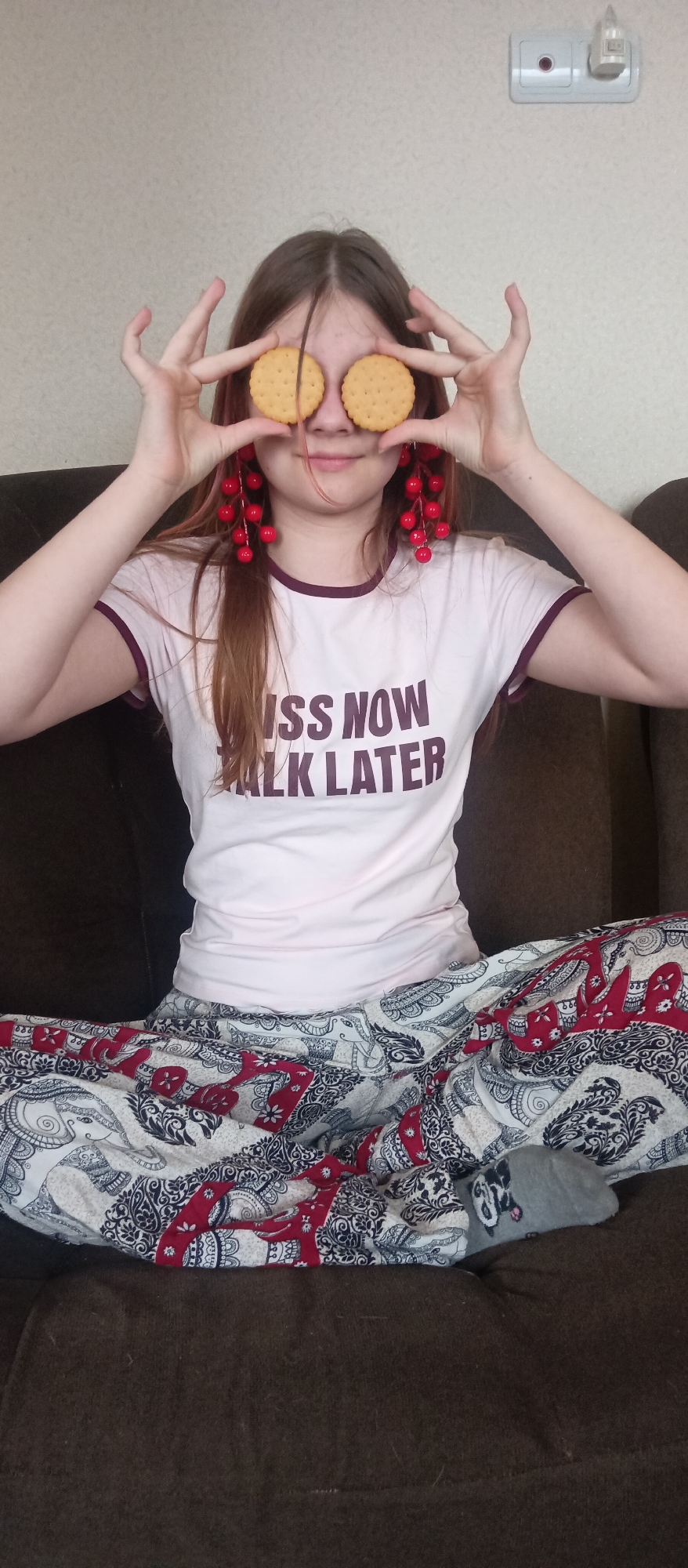 A young person with long brown hair is seated cross-legged on a dark brown upholstered couch, playfully holding two round, light-colored biscuits over their eyes. Their fingers are splayed around the biscuits, framing them. A slight smile is visible on their face, suggesting a lighthearted or amusing moment.
The person wears a light pink or off-white t-shirt with maroon trim around the collar and short sleeves. Bold text on the front of the shirt reads "KISS NOW" and "ALK LATER," implying the phrase "KISS NOW TALK LATER," with the initial 'T' of 'TALK' obscured by their arm. Prominent, long, dangling red earrings, resembling clusters of berries, adorn their ears.
They are dressed in patterned pajama-style pants featuring an intricate design of elephants, paisley motifs, and floral patterns in white, red, and dark blue/black. A single gray sock with a subtle dark pattern is visible on their left foot.
The background shows a plain, light-colored wall. In the upper right corner, a white electrical outlet with a plugged-in white device (possibly a night light or air freshener) is visible. The setting appears to be a casual indoor environment, likely a home living space, during an undeterminable time of day. The overall mood is cheerful and relaxed.