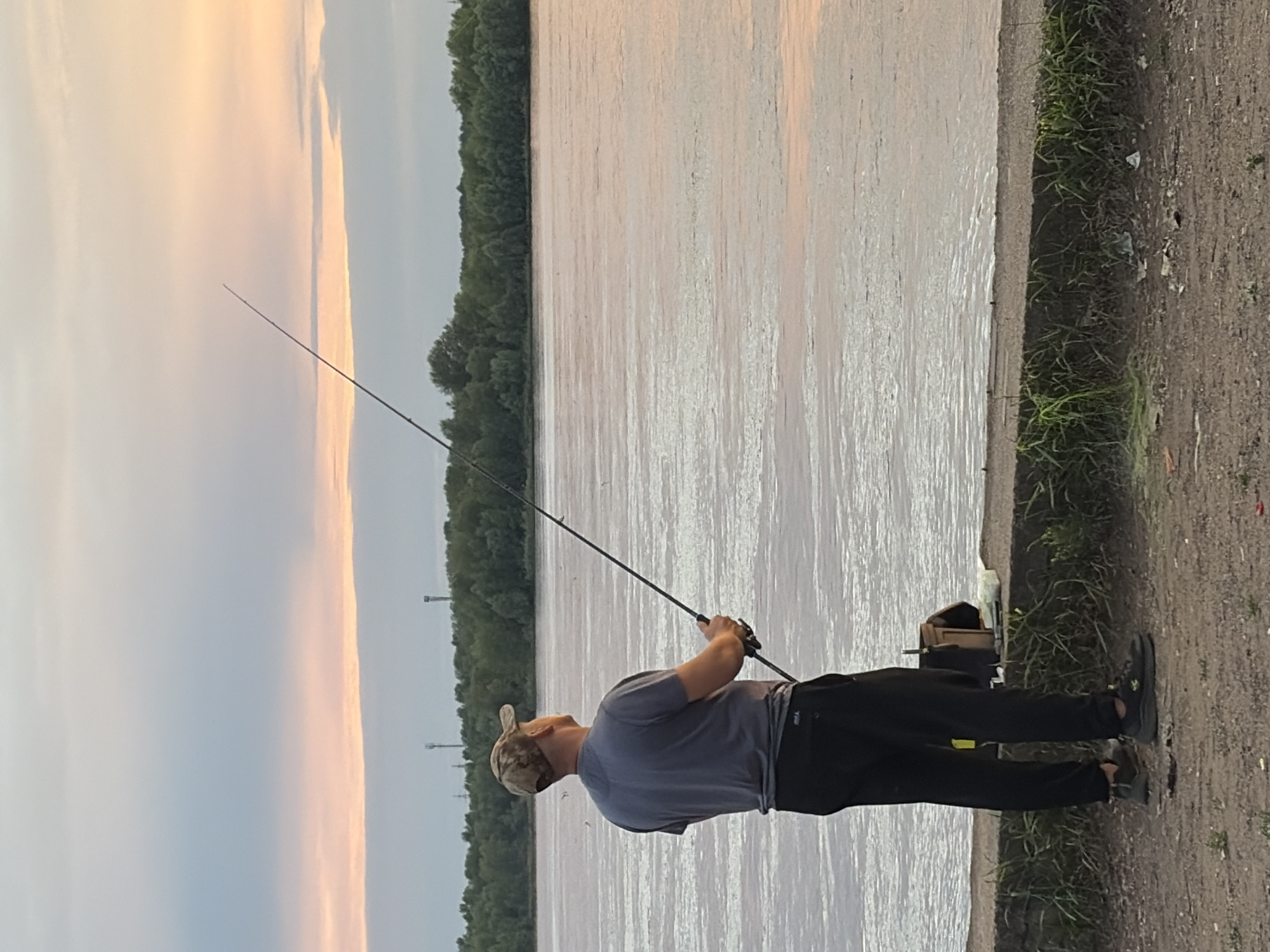 A man wearing a camouflage baseball cap, a grey t-shirt, and black pants stands facing away from the camera, holding a fishing rod over a wide body of water. The water is calm with a slight ripple effect, reflecting the muted colors of the sky. In the background, a dense line of green trees borders the opposite bank of the river. The sky above is a gradient of soft pink and pale blue, indicative of either sunrise or sunset. The man is positioned on a concrete embankment, with a small tackle box visible beside him. The ground in the foreground is a mix of dirt and sparse green grass, with some scattered litter. The overall scene is one of quiet solitude and outdoor recreation along the Kama River in Chaikovsky, Russia.