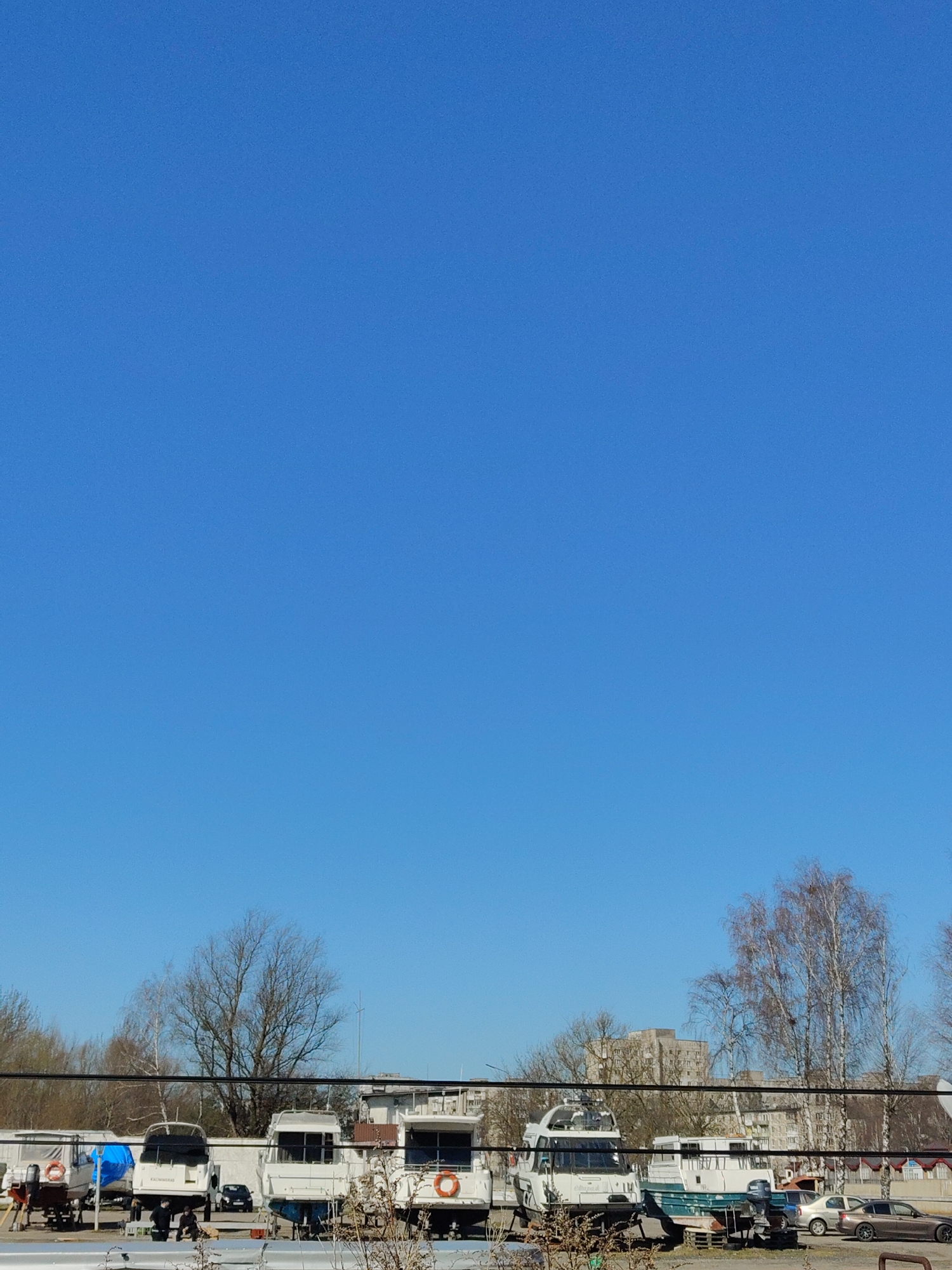 The image shows an outdoor scene in a location identified as "Unknown, Unknown," likely a storage or maintenance yard. The sky is clear and blue, filling most of the frame. A row of boats on trailers occupies the lower portion of the image. The boats vary in size and design, with some covered in tarpaulins. Buildings are visible in the background behind the boats. Trees with bare branches are scattered throughout the scene. Power lines run across the lower third of the image, slightly obscuring the boats and buildings. Several vehicles are parked nearby. Some people are on the ground near the boats.