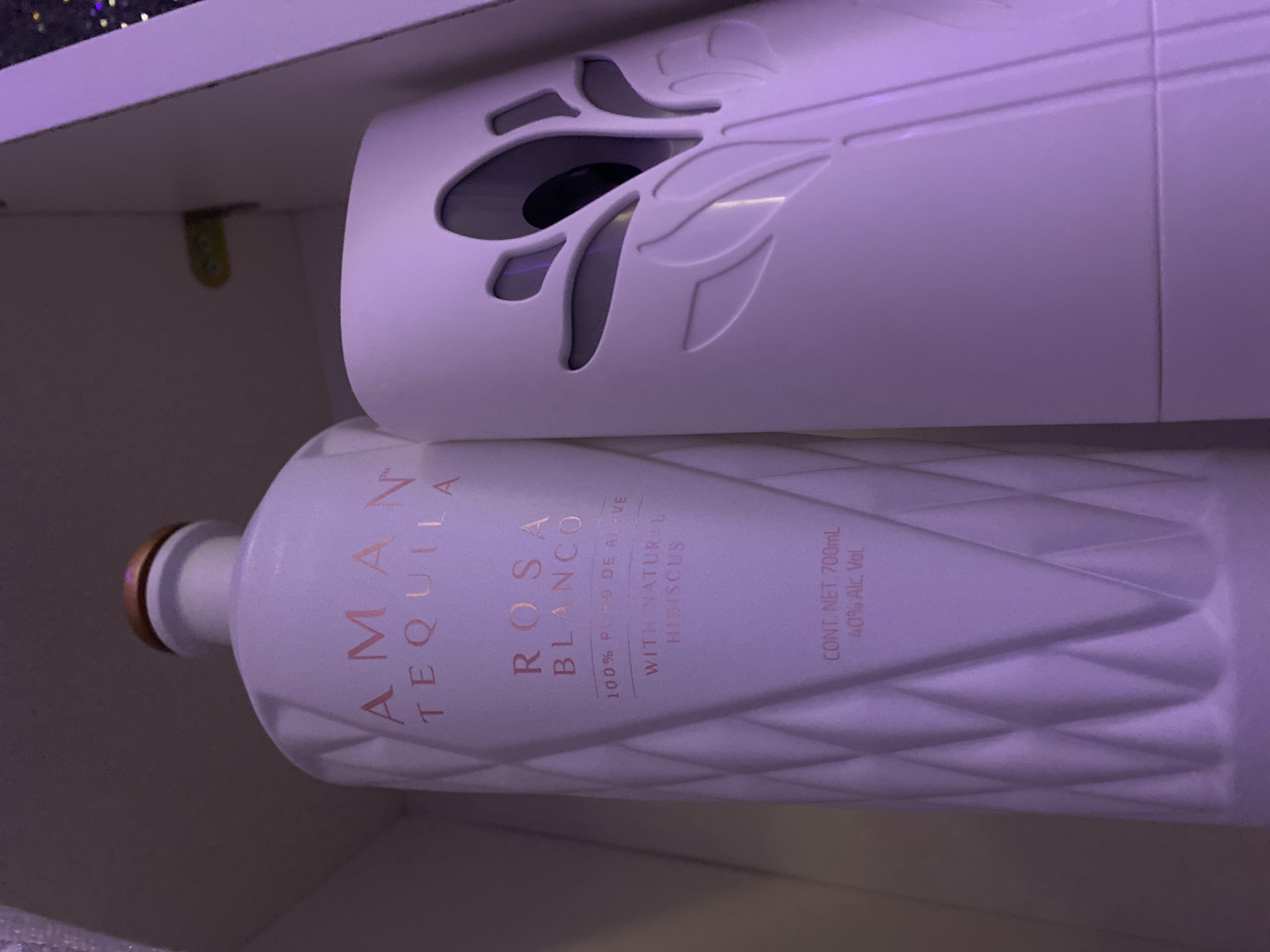 A vertically oriented, close-up shot captures a white bottle of "AMÁN TEQUILA ROSA BLANCO" and a white air freshener with a floral cutout design. The bottle, with a gold cap, is positioned on the left side of the frame and features rose gold lettering detailing its contents: "100% PURO DE AGAVE WITH NATURAL HIBISCUS CONT. NET. 700mL 40% Alc. Vol." The air freshener, to the right of the bottle, has an intricate floral pattern on its front, revealing a dark interior. The objects are situated within a white, enclosed space, possibly a shelf or cabinet, illuminated by a purple light that casts a soft glow over the scene. The background is out of focus and appears to be a plain, dark surface. There are no people present in the image. The location in Kason, Nigeria, is not determinable from the visual information provided.