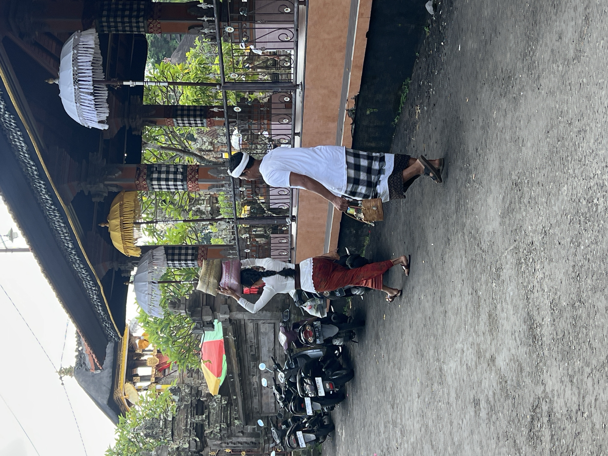 The image depicts a street scene in Singakerta, Indonesia. A man and a woman are carrying offerings, with the woman holding baskets on her head. The man is wearing a white shirt, a checkered sarong, and a white headband; he carries a woven container. The woman wears a white blouse and a red sarong. Several motorcycles are parked nearby. The scene is set against a backdrop of a building decorated with traditional Balinese umbrellas and checkered cloths. The ground is a gray paved surface.