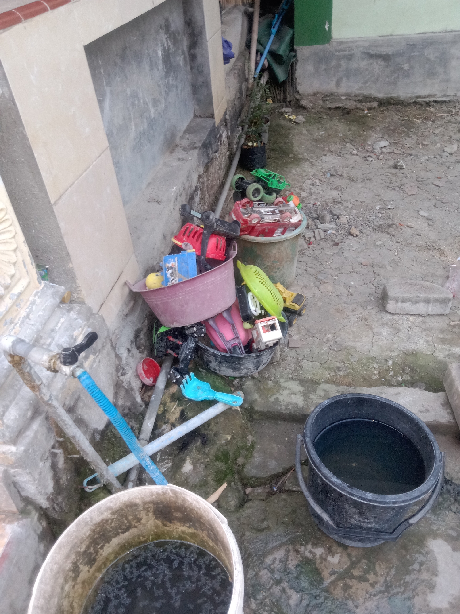 The image depicts an outdoor scene with various children's toys and containers in a state of disarray. The location is identified as Jumput, Indonesia. In the foreground, two large buckets are visible. The one on the left is partially filled with dark, murky water. The bucket on the right is filled with water and appears to be a black plastic pail.
Surrounding these buckets, an assortment of toys, primarily toy trucks and cars, are scattered around. Some of the toys are contained within a large pink basin, which also holds a yellow ball and a blue rectangular object. Other toys are loose on the ground, including a green toy truck, a red toy truck, and a yellow toy excavator. A blue plastic rake is also present.
The environment appears to be an outdoor area with a concrete or dirt ground surface, which is cracked in places and appears damp. A wall constructed of concrete blocks and possibly stone is visible on the left side of the image, with a water tap and a blue hose attached to it. In the background, there is a building with a light-colored wall and a green section. Vegetation, in the form of a small potted plant, is also visible. The overall impression is of a cluttered outdoor space where children's toys have been left.