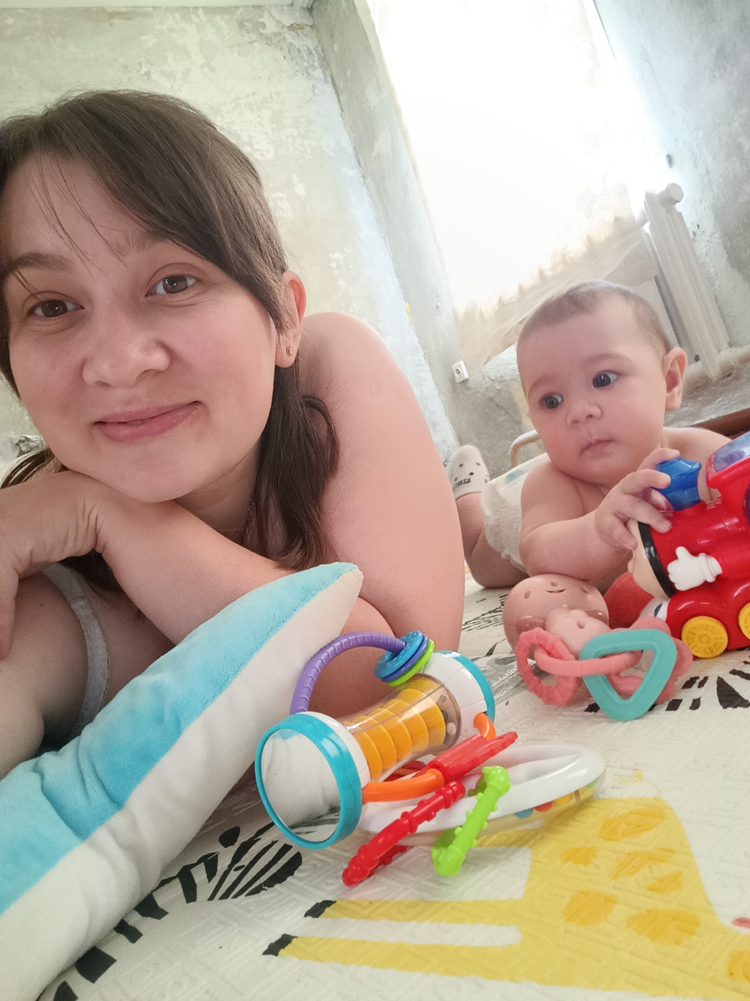 A woman and a baby are lying on their stomachs on a patterned blanket on the floor. The woman is in the foreground, looking at the camera with a slight smile and her chin resting on her hand. She has dark hair and is wearing a light-colored tank top. Beside her is a blue and white plush pillow. The baby is to her right, looking off to the side with a curious expression. The baby is wearing a diaper and white socks with black stripes. In front of the baby are several colorful baby toys, including a red toy train, a rattle with rings, and a clear plastic toy with colorful beads inside. The background appears to be an indoor setting with a light-colored wall and a radiator. Based on the provided information, the location is specified as Khokhly, Russia.